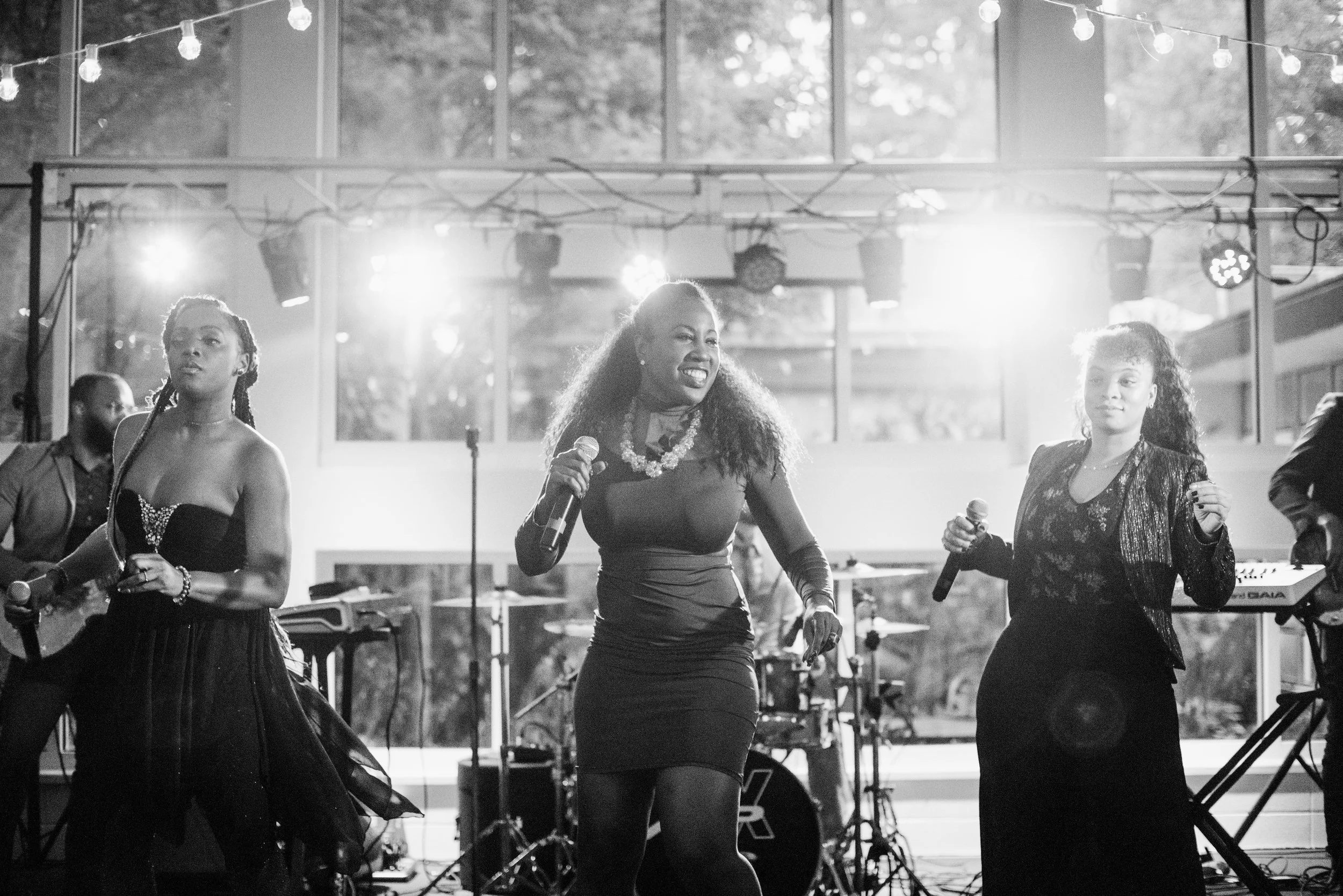 Women singing and playing musical instruments during an outdoor performance, with stage lighting and sound equipment visible.