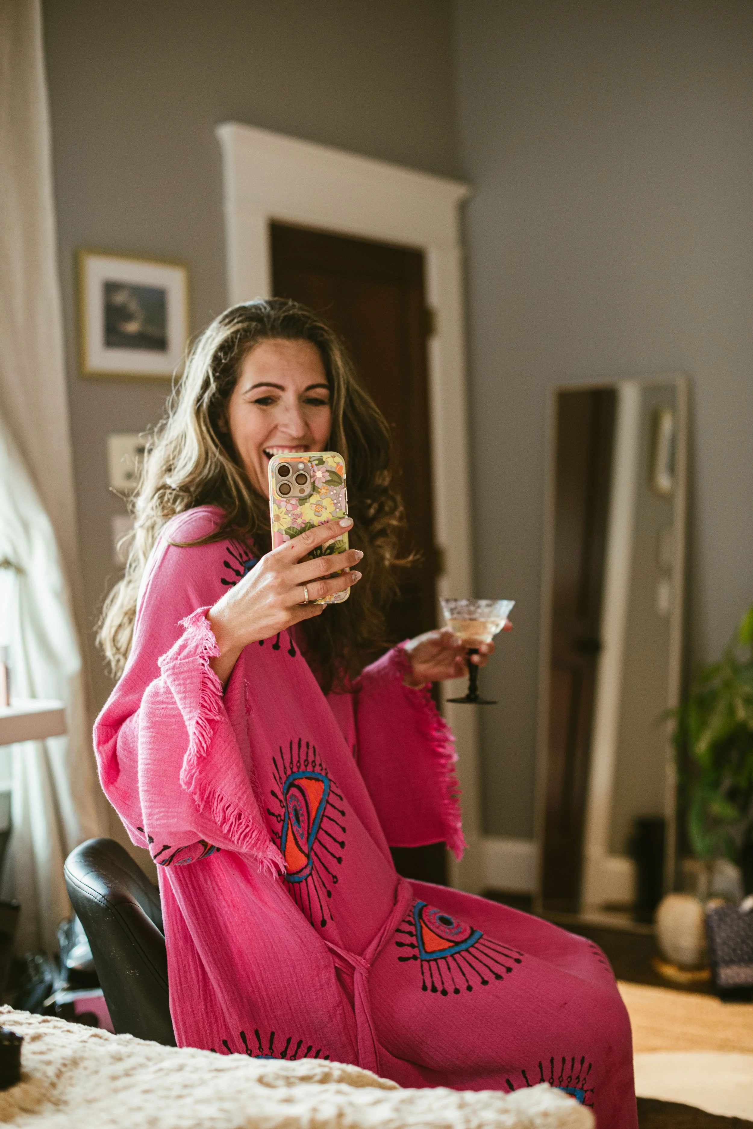 A woman with long, curly hair in a pink dress with colorful designs on it, is smiling and taking a selfie in a mirror while holding a glass of wine.
