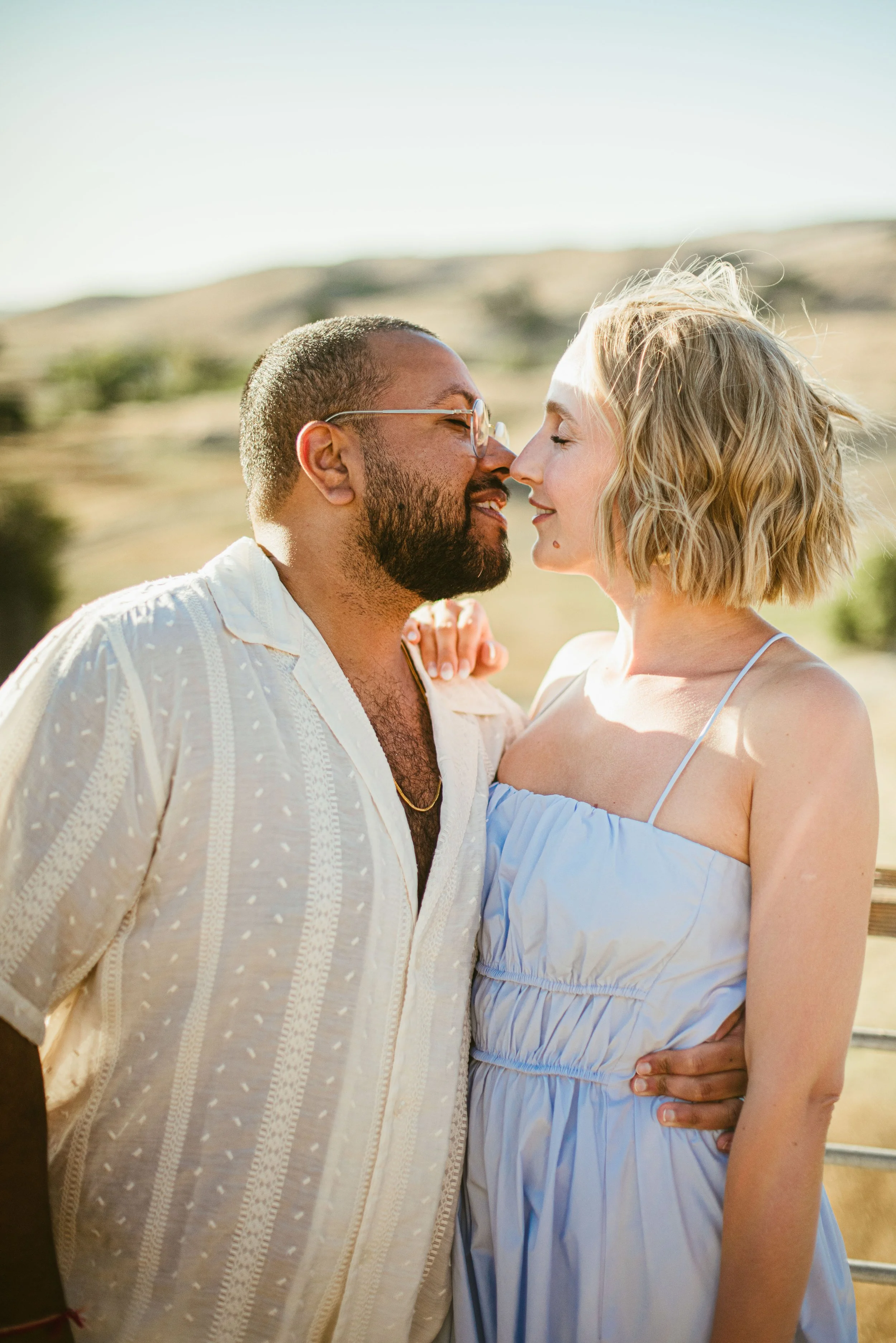 A couple standing close together outdoors with their foreheads touching, smiling, and looking into each other's eyes. The man has a beard, glasses, and is wearing a cream-colored shirt, while the woman has short blonde hair and is wearing a light blu