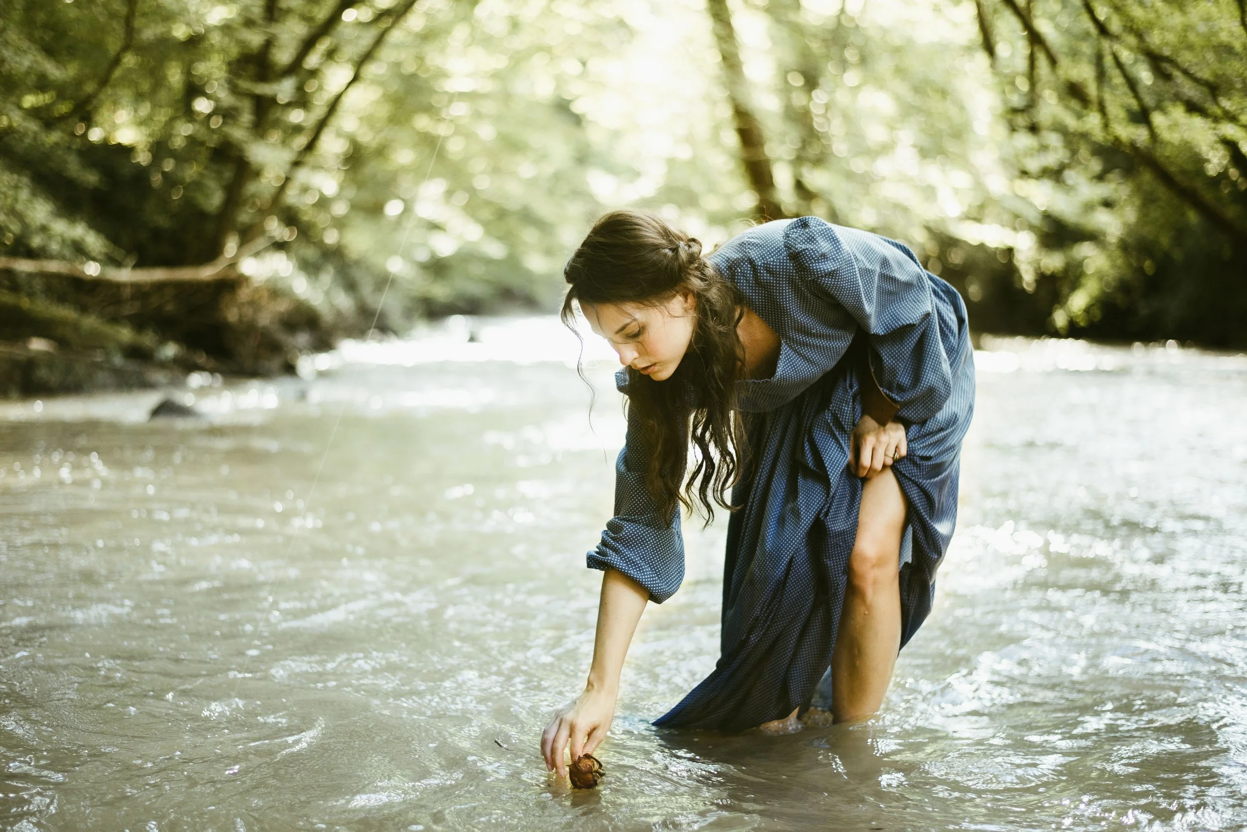 A woman with long dark hair, wearing a blue dress, is in a shallow river, bent over reaching into the water, with green trees and sunlight in the background.