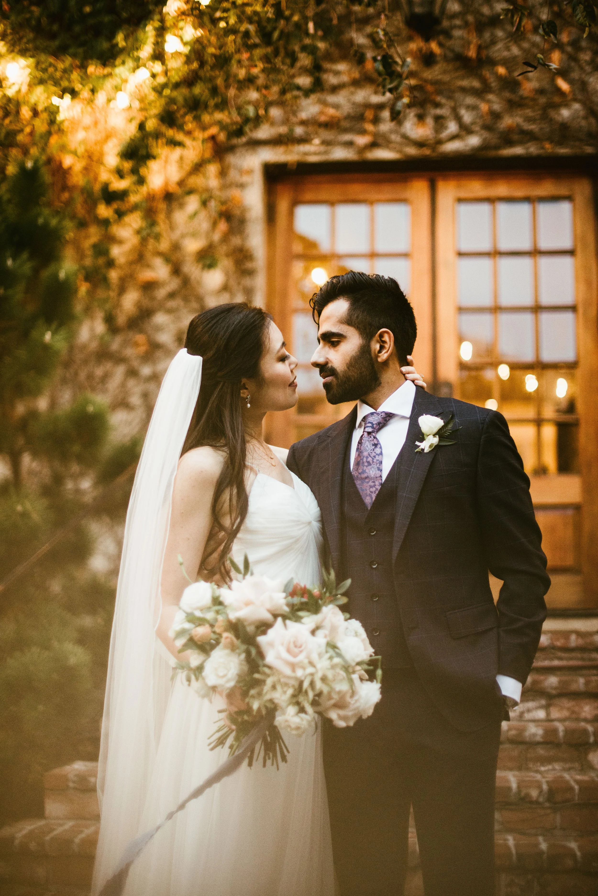 A bride and groom are facing each other closely during their wedding. The bride is holding a bouquet of white and blush flowers, with a long white veil. They are standing outside in front of a rustic brick building with warm lighting, surrounded by g