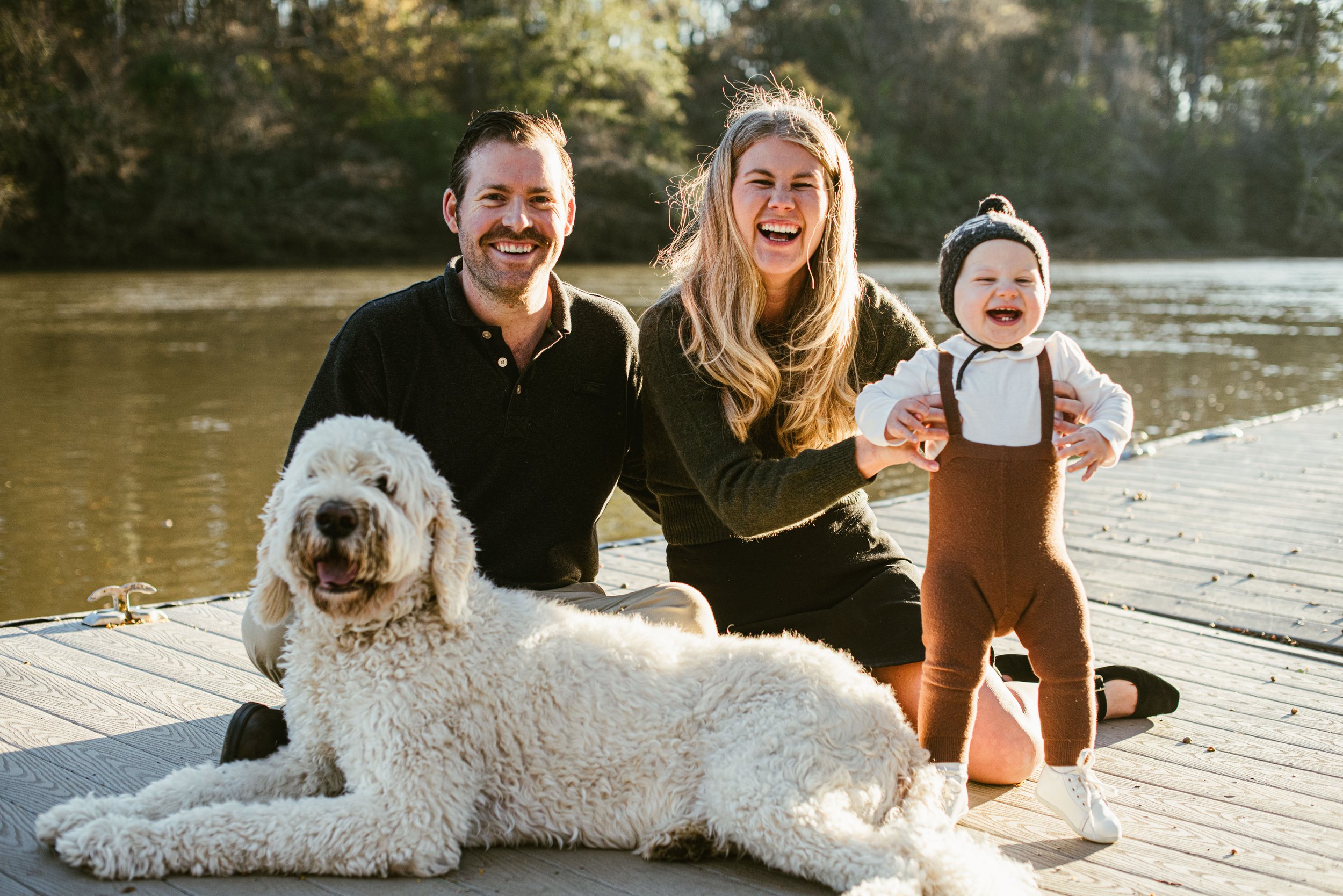 Family of three, a man, woman, and a young child, sitting on a dock by a river with their dog, enjoying a sunny day outdoors.