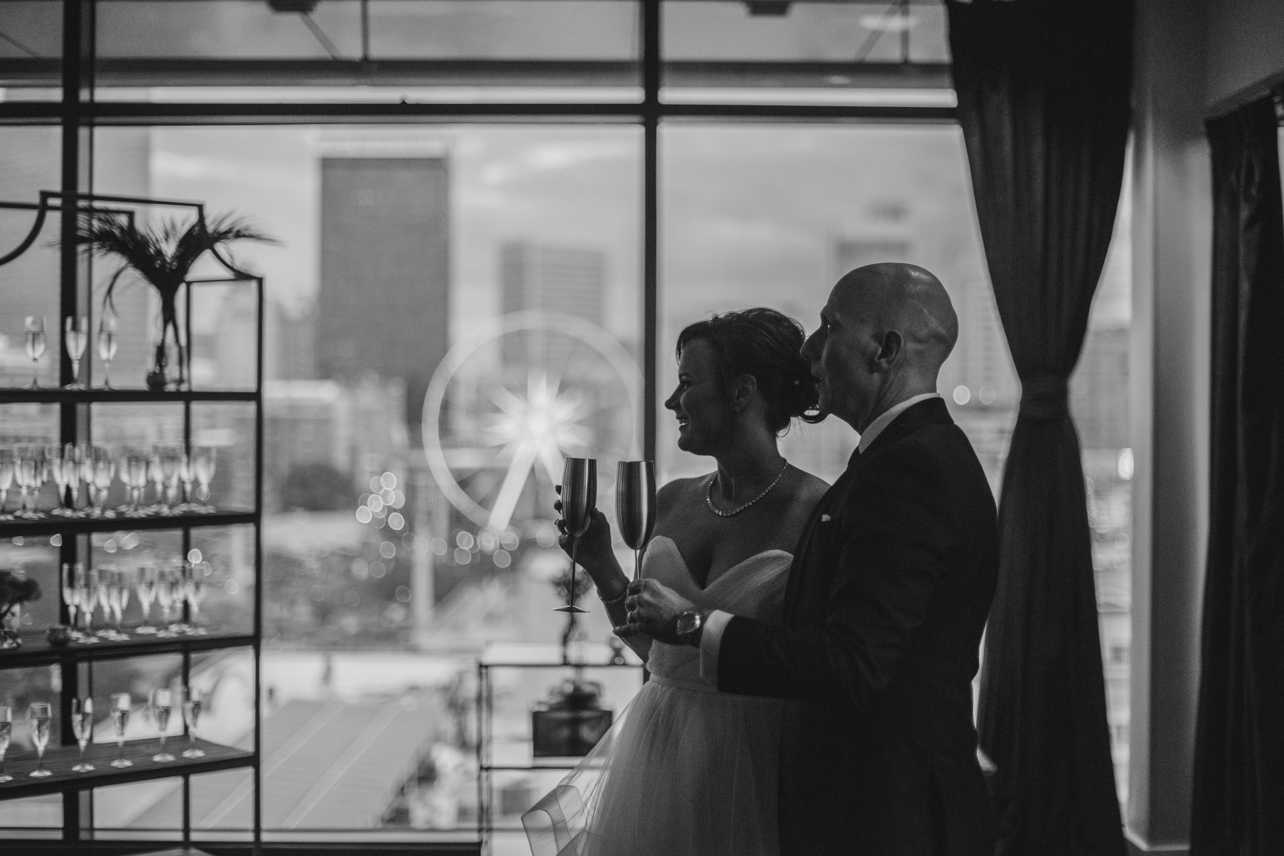 A black and white photo of a smiling woman and a man in formal attire holding glasses of champagne, standing near a window during what appears to be a celebration or wedding. The background shows a cityscape and a Ferris wheel.