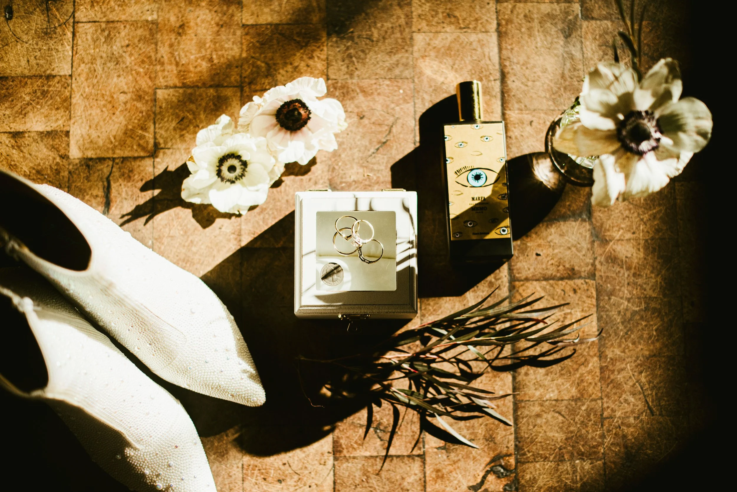 A top-down view of flowers, jewelry, perfume, and a shoe on a brick floor with shadows.