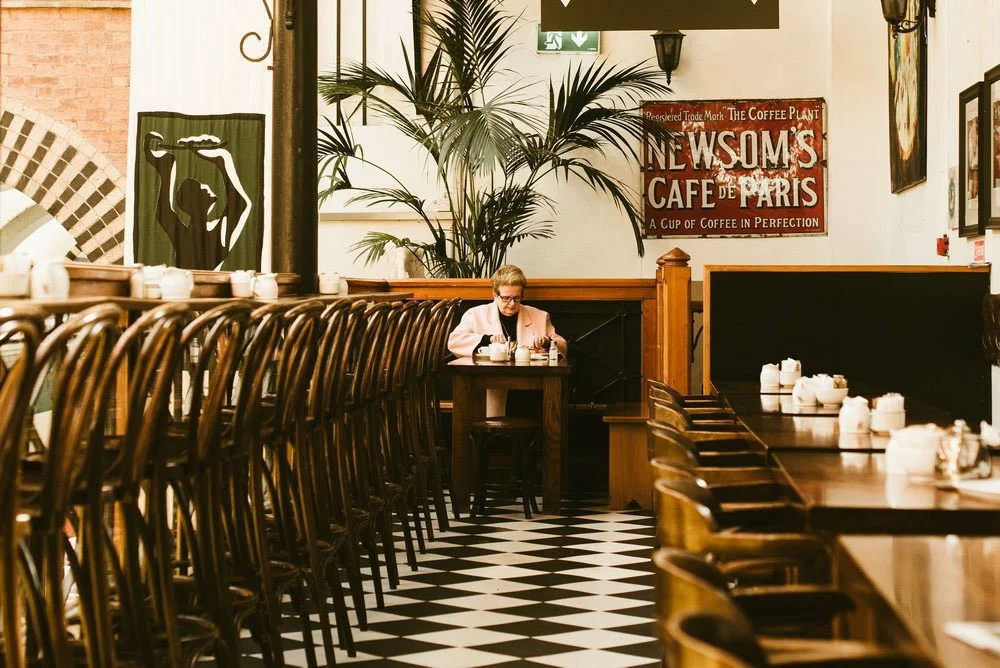 A mostly empty cafe with wooden chairs and tables, a woman sitting alone at a table, large plants, and vintage signs on the wall.