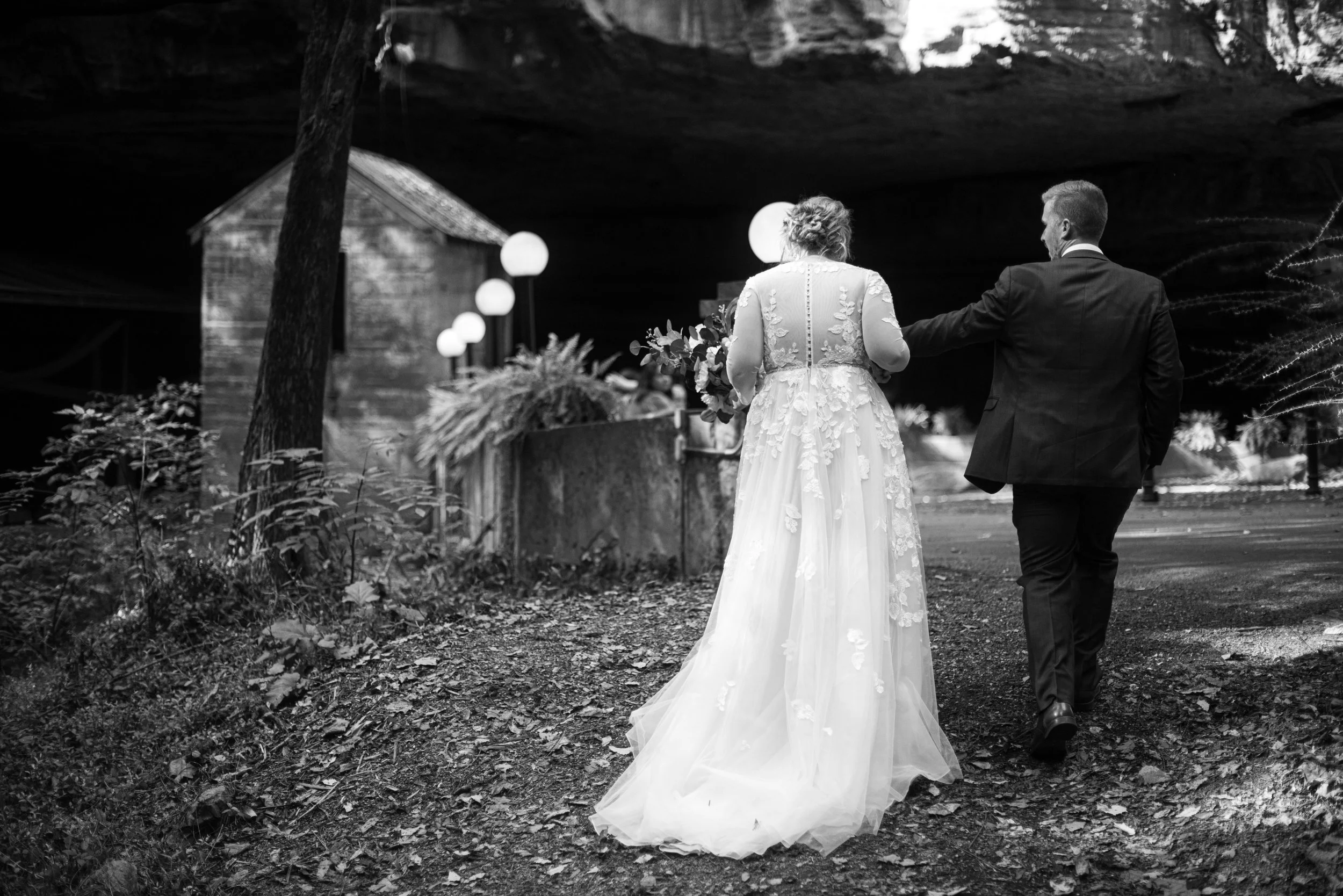 Black and white photo of a bride and groom walking under a large rock overhang, with trees and foliage around them. The bride is in a lace wedding dress holding a bouquet, and the groom is in a suit.