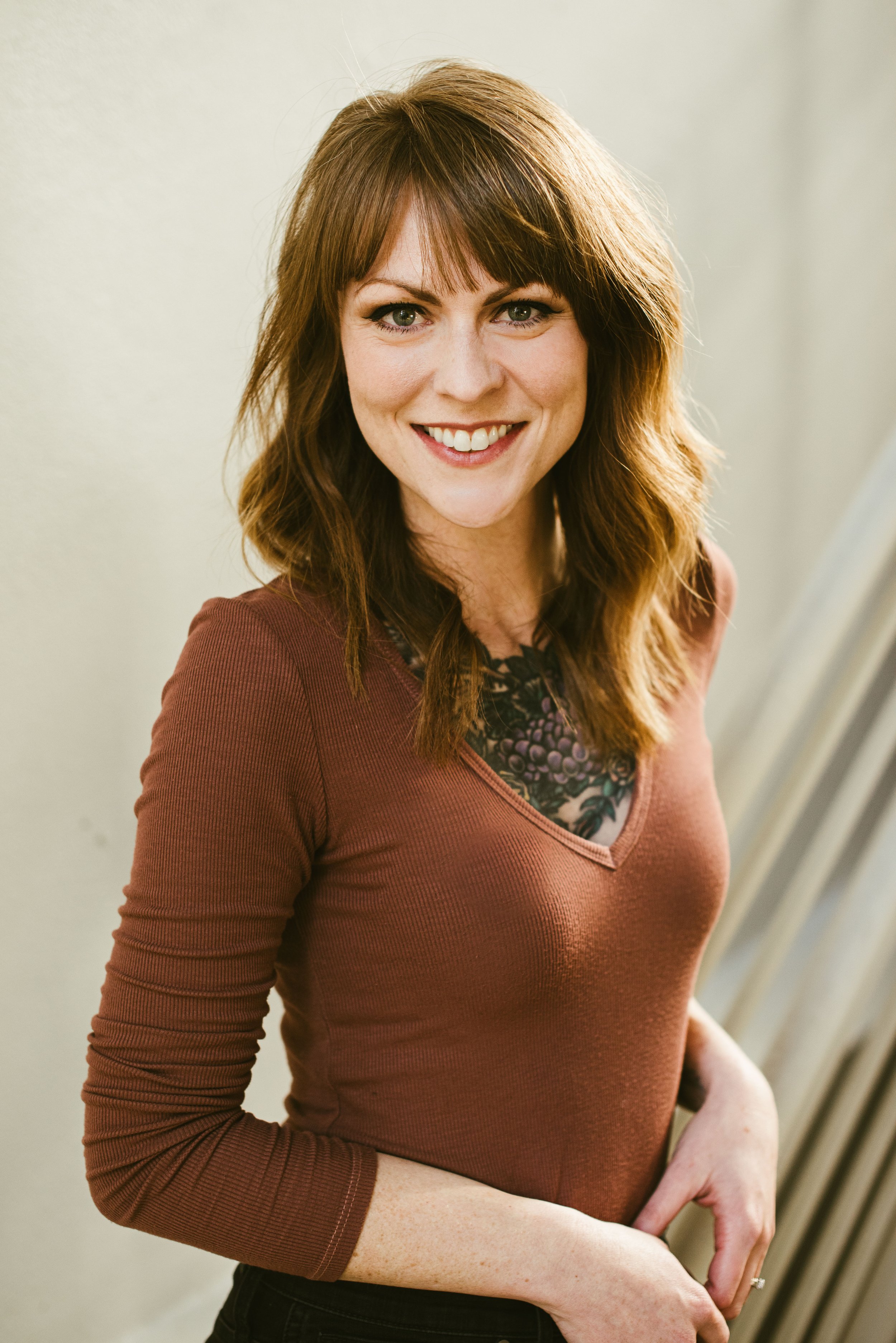A woman with reddish-brown hair, wearing a brown top and a necklace with purple beads, smiling and posing for the photo. She is standing against a plain light background.