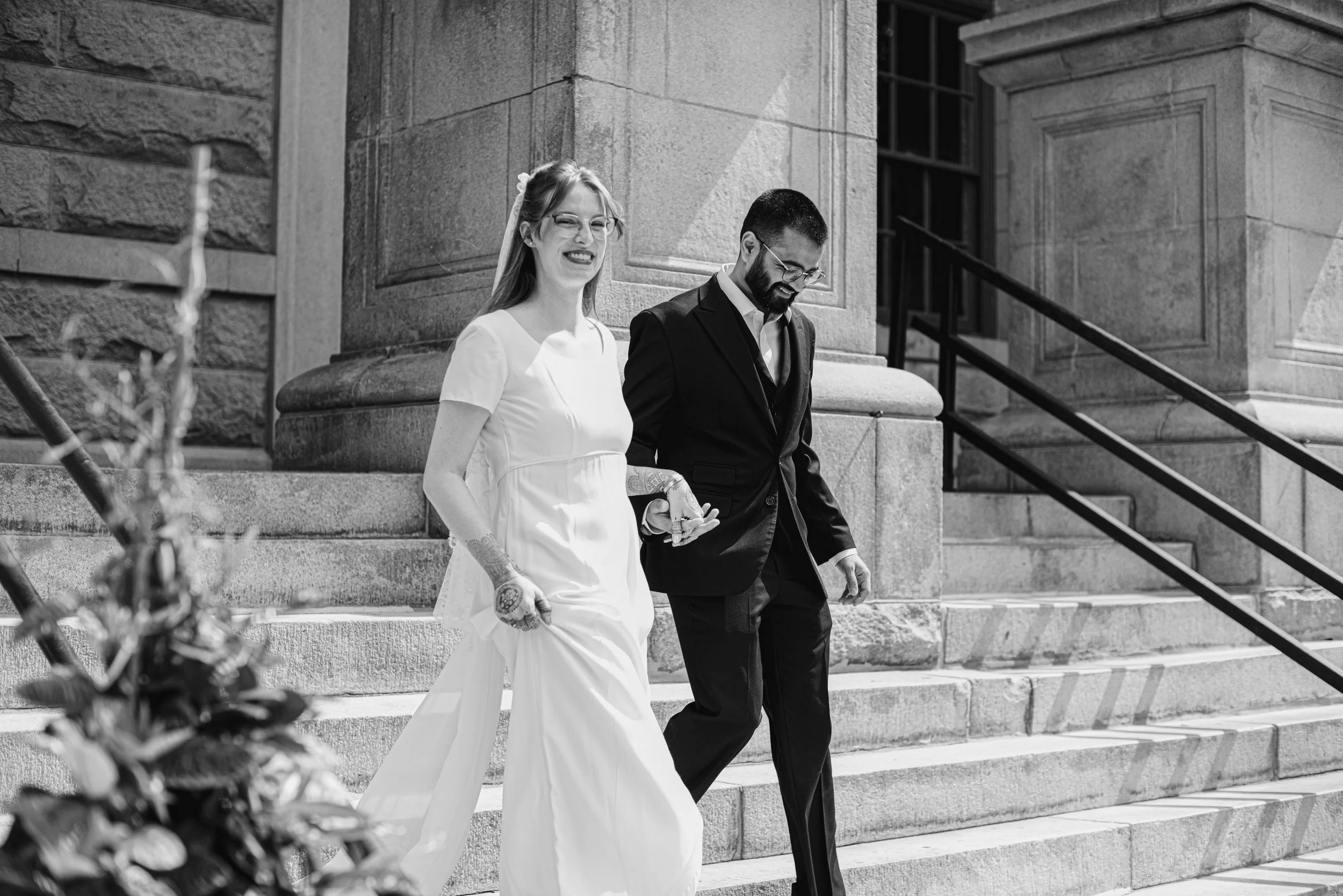 A black and white photo of a bride and groom walking down the steps outside a stone building, smiling and holding hands.