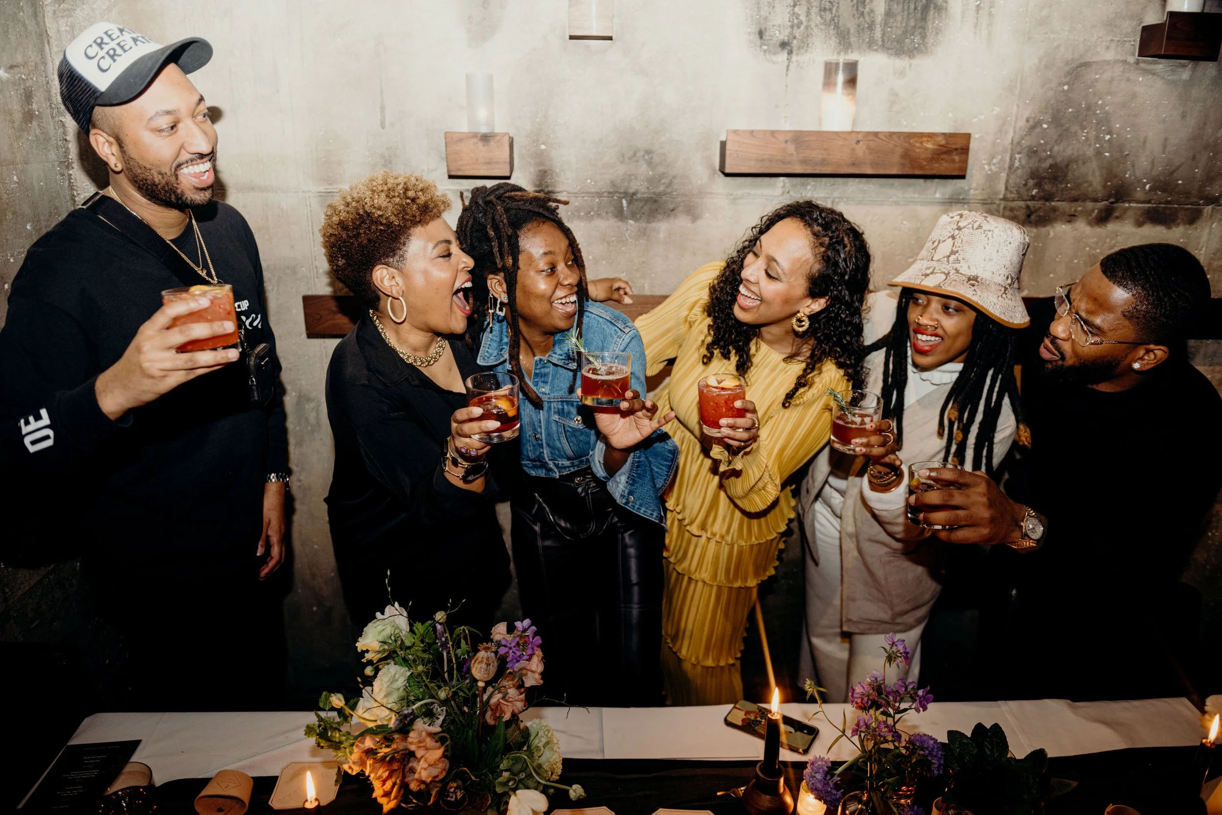A group of seven people gathered around a table with flowers and candles, celebrating with drinks, smiling and laughing together in an indoor setting with a concrete wall background.