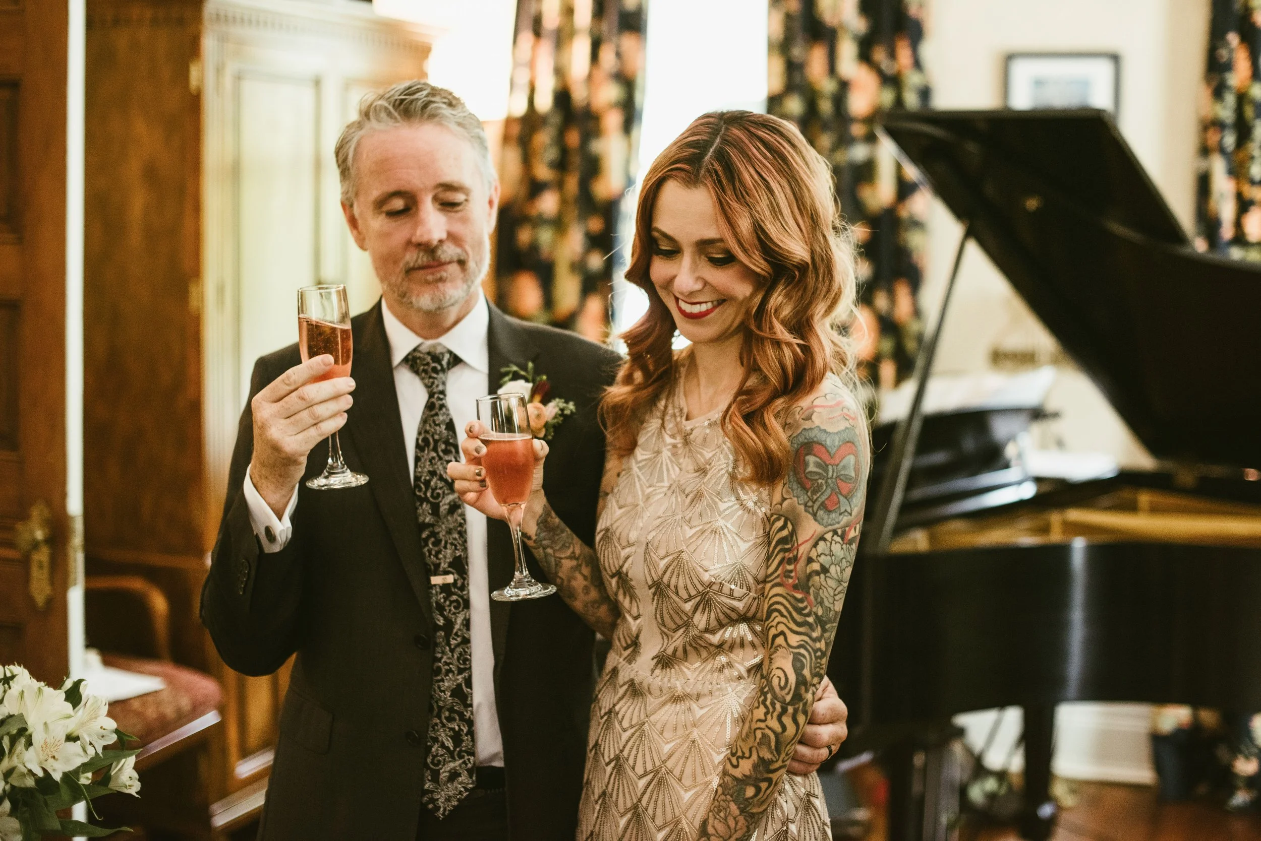 A man in a dark suit and a woman with long red hair and tattoos on her arms, both holding glasses of pink champagne, are smiling and looking at each other during a celebration, with a grand piano in the background.