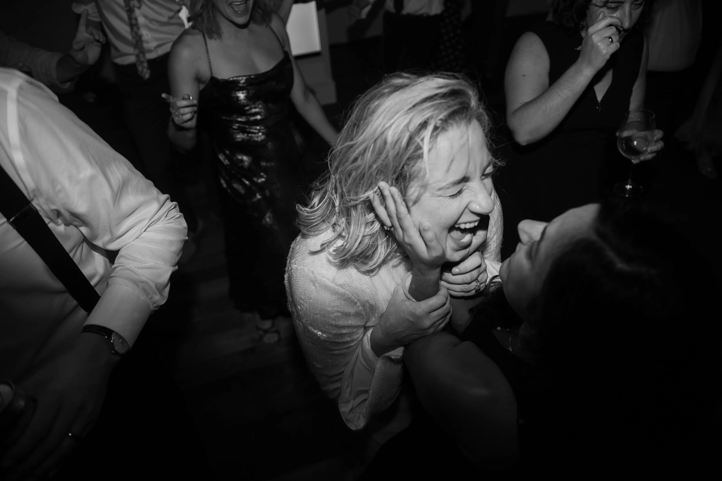 A woman with curly hair laughing joyfully at a social gathering, surrounded by other people holding drinks in a black-and-white photo.