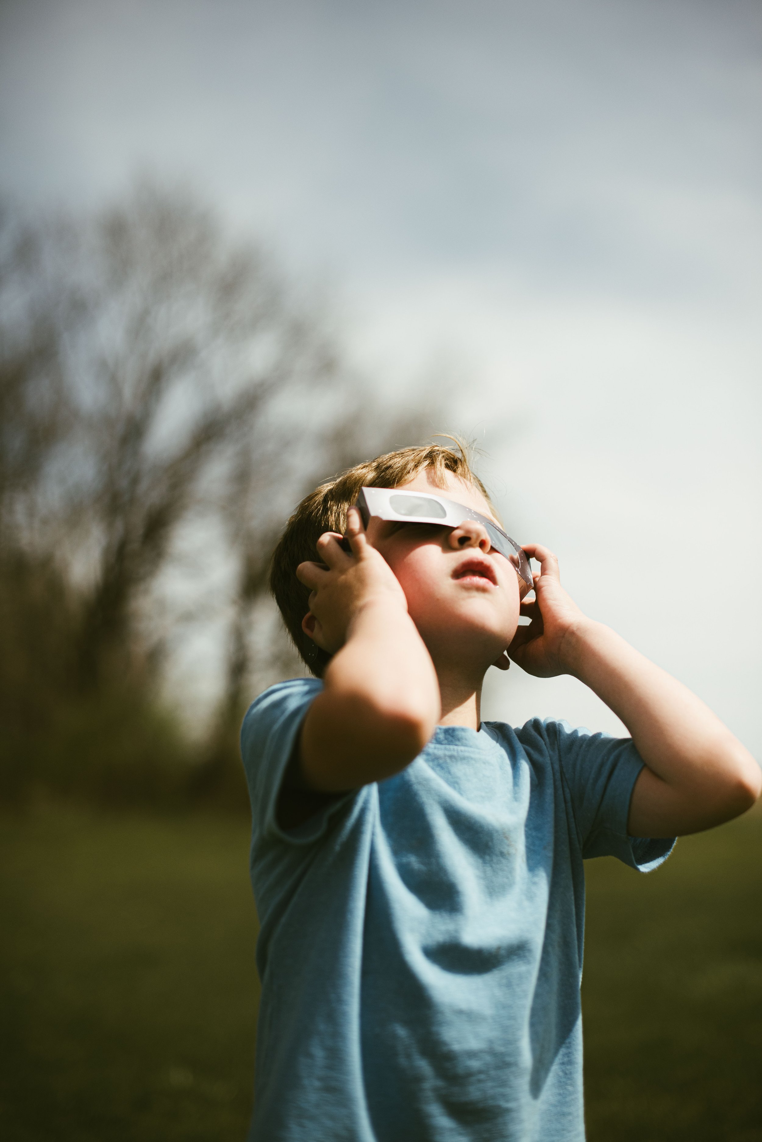 A young boy wearing sunglasses and a blue t-shirt is outdoors, adjusting his sunglasses with both hands, with blurred trees and sky in the background.