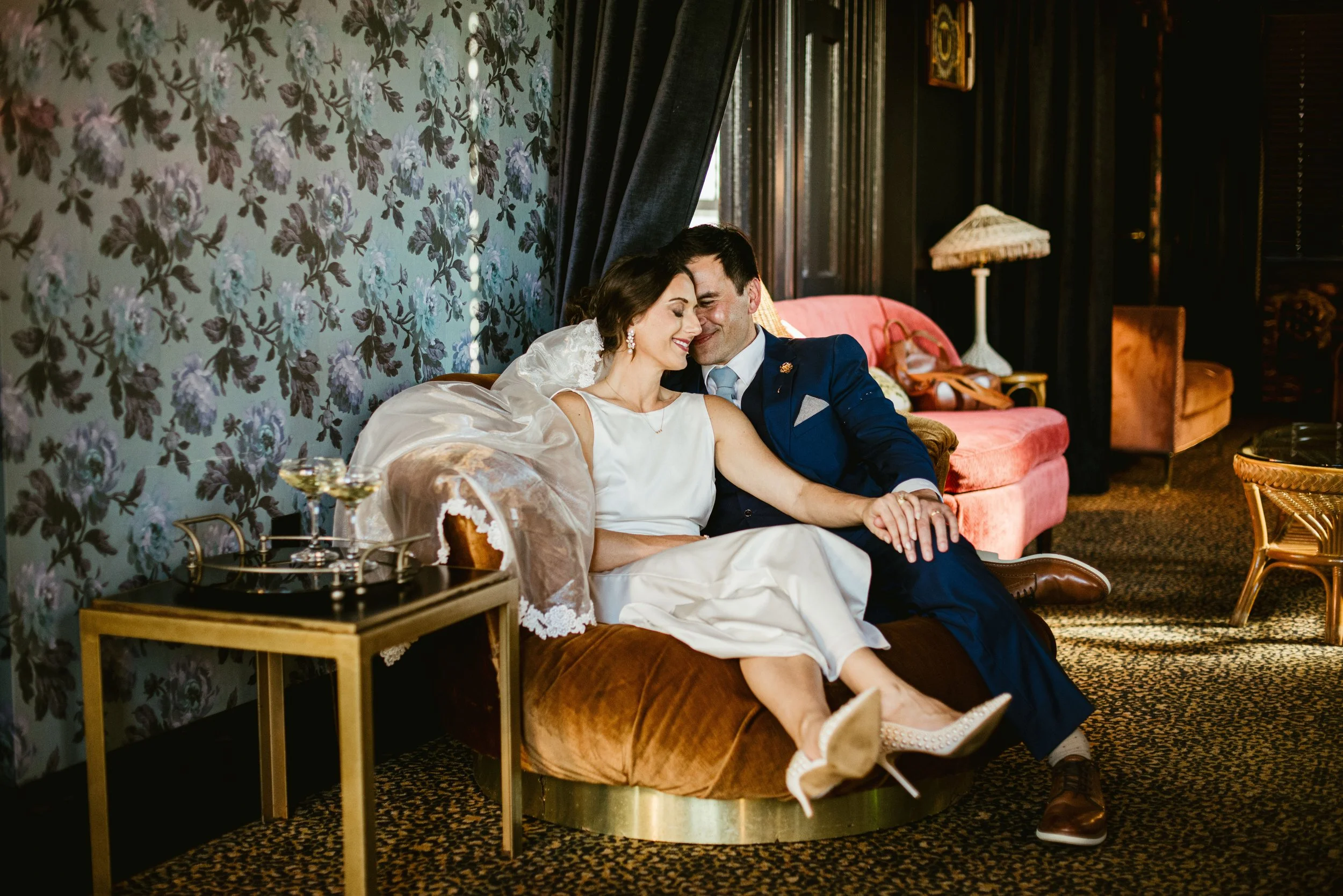 A bride and groom sitting close together on a vintage orange velvet sofa, smiling and sharing an intimate moment in a cozy, dimly lit room decorated with floral wallpaper, dark curtains, a pink armchair, and a lamp in the background.