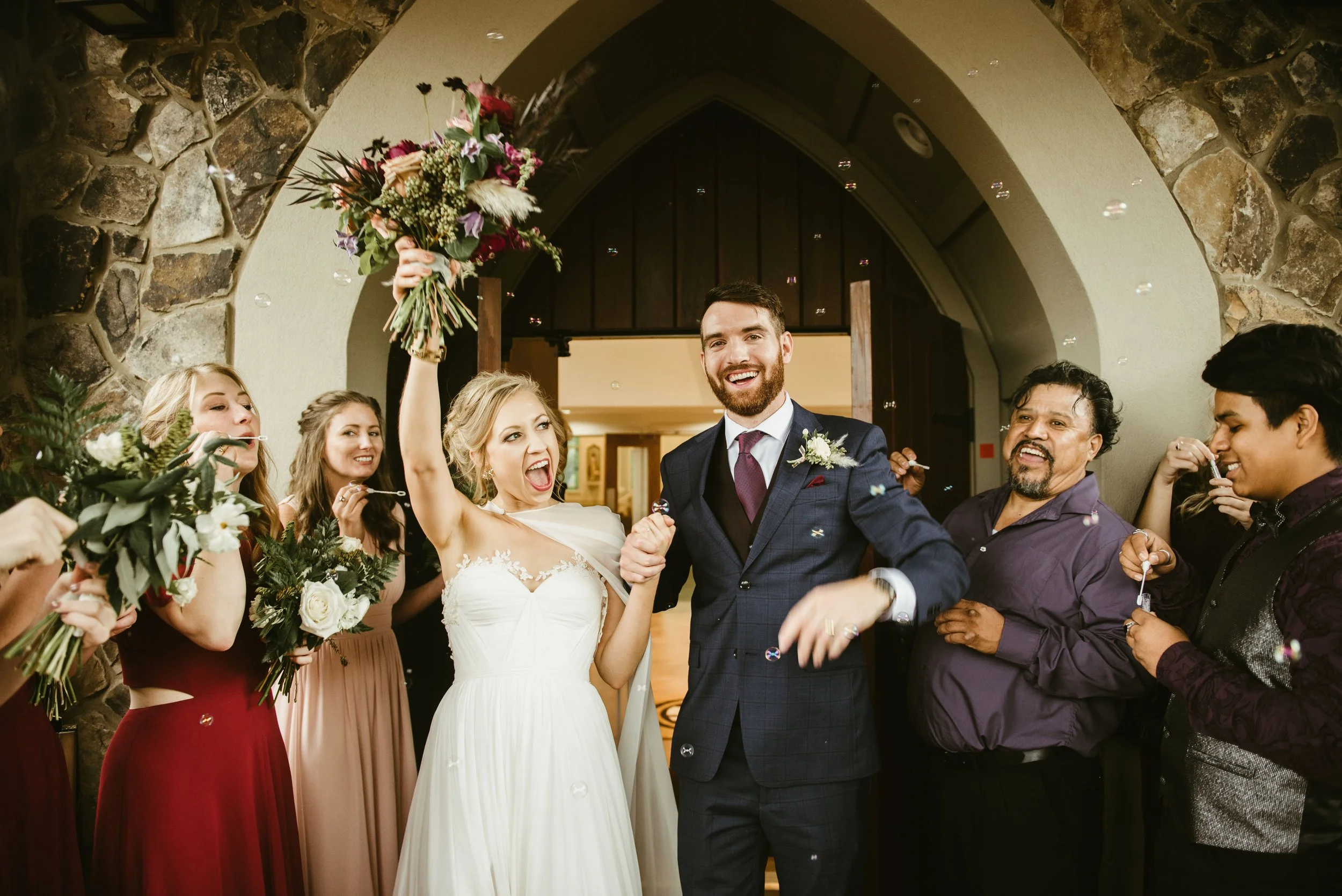 A newlywed couple celebrating, surrounded by friends. The bride is holding a bouquet and the groom is dressed in a suit, all smiling and joyfully throwing confetti.