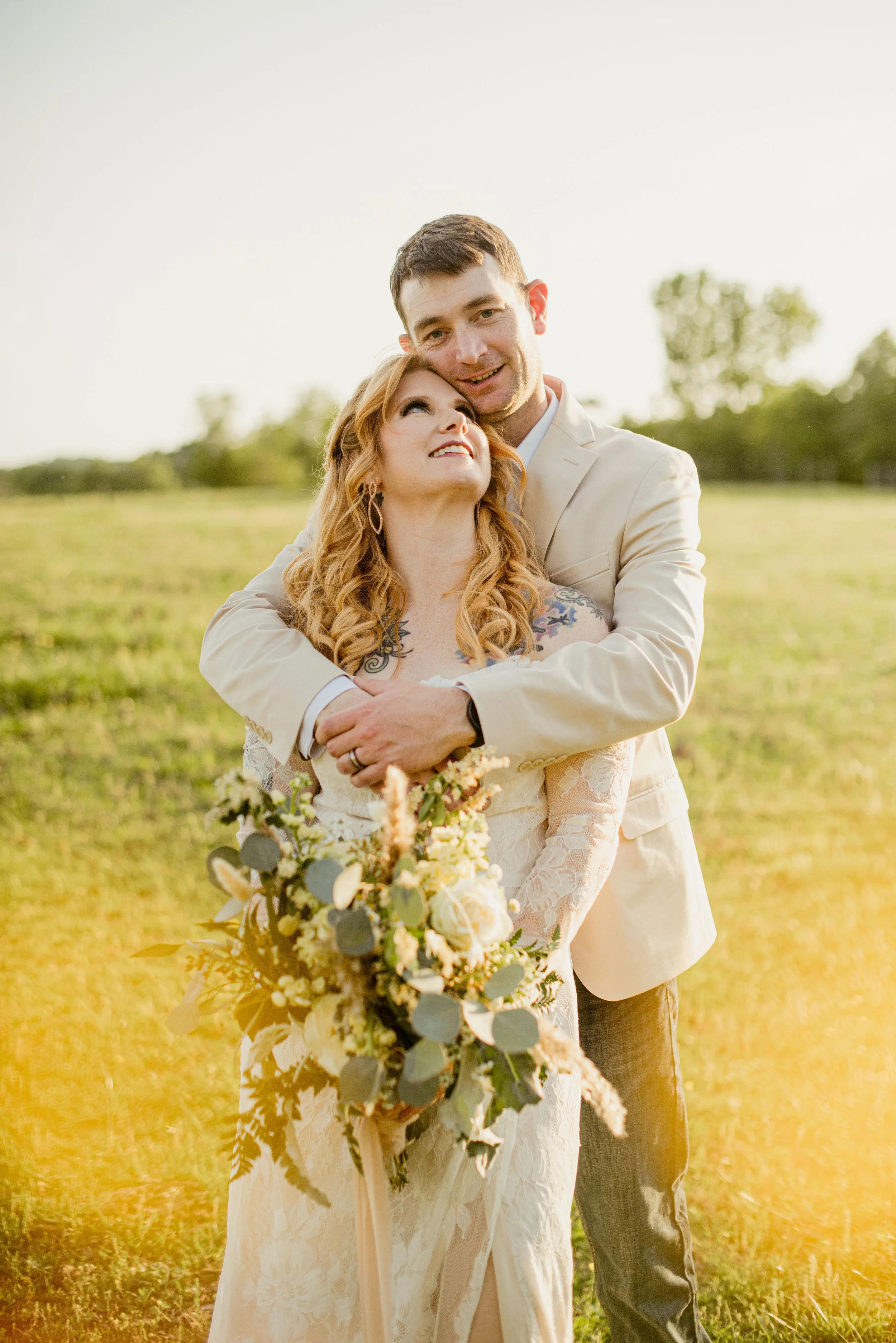 A newlywed couple in wedding attire embracing outdoors in a grassy field with trees in the background during sunset.