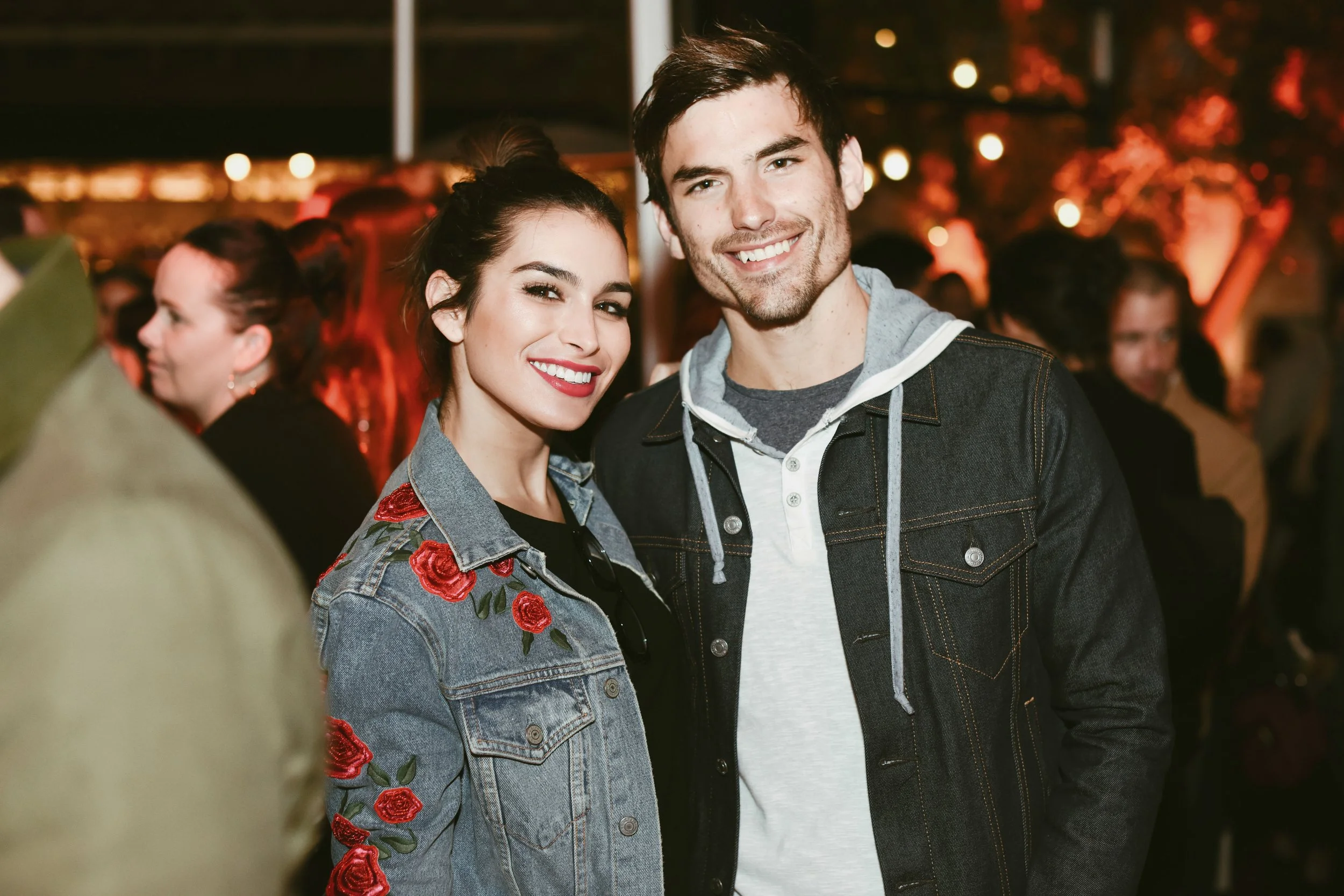 Jared and Ashley at a social event, with blurred people and warm lighting in the background.
