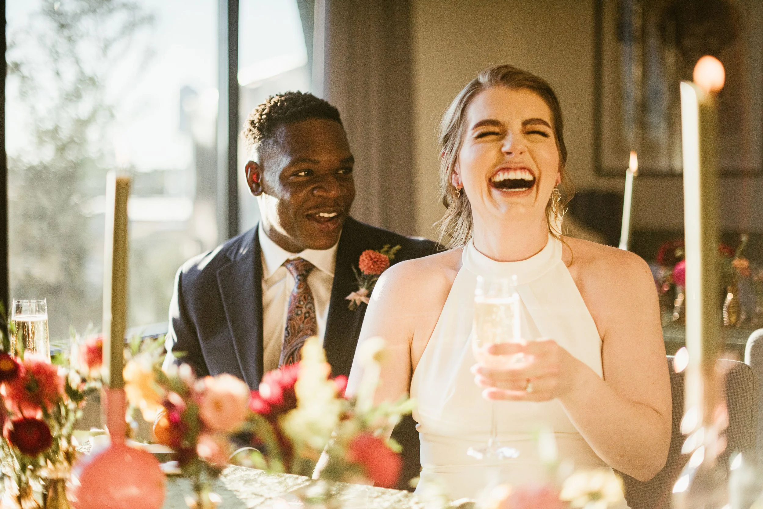 A young woman in a white dress laughing and holding a glass of champagne during a celebration, with a man in a suit smiling behind her, sitting at a decorated table with flowers and candles, in a warmly lit room with a window.