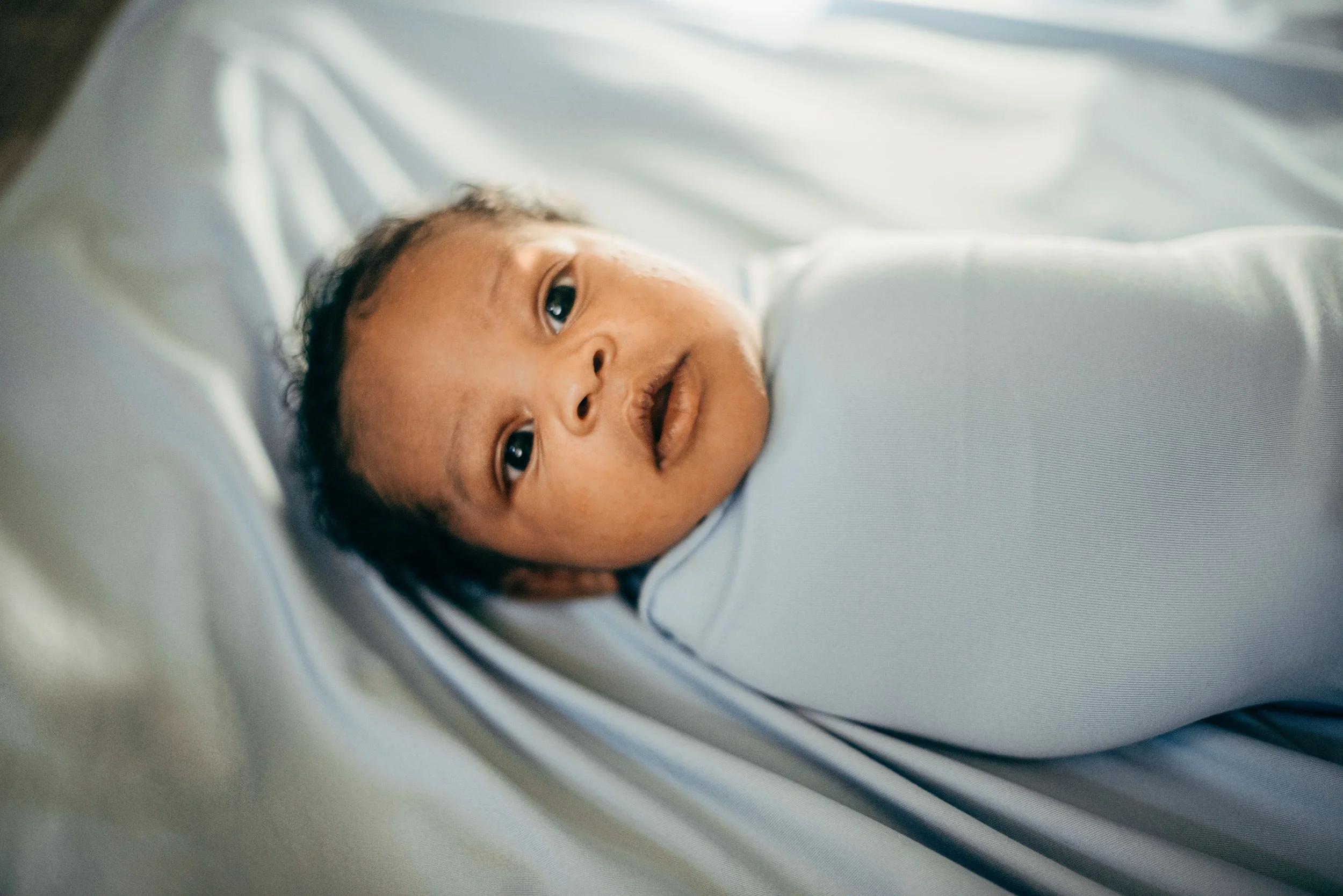 Close-up of a baby's face lying on a white surface, looking towards the camera with dark eyes.