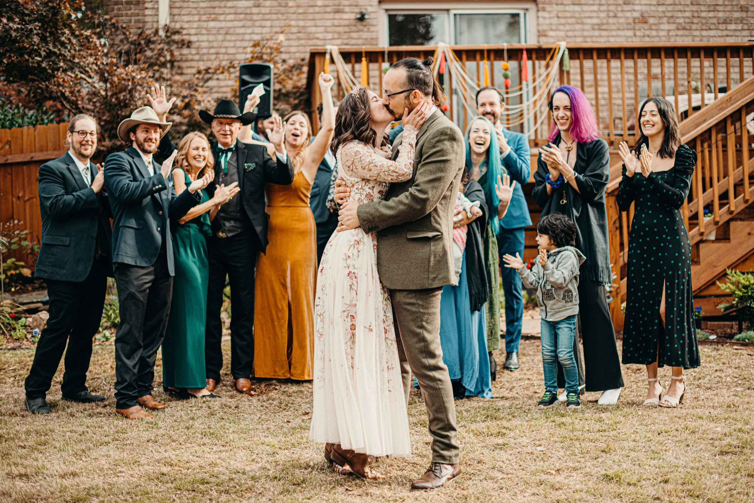 Couple kissing at outdoor wedding reception with friends clapping and cheering in backyard setting.