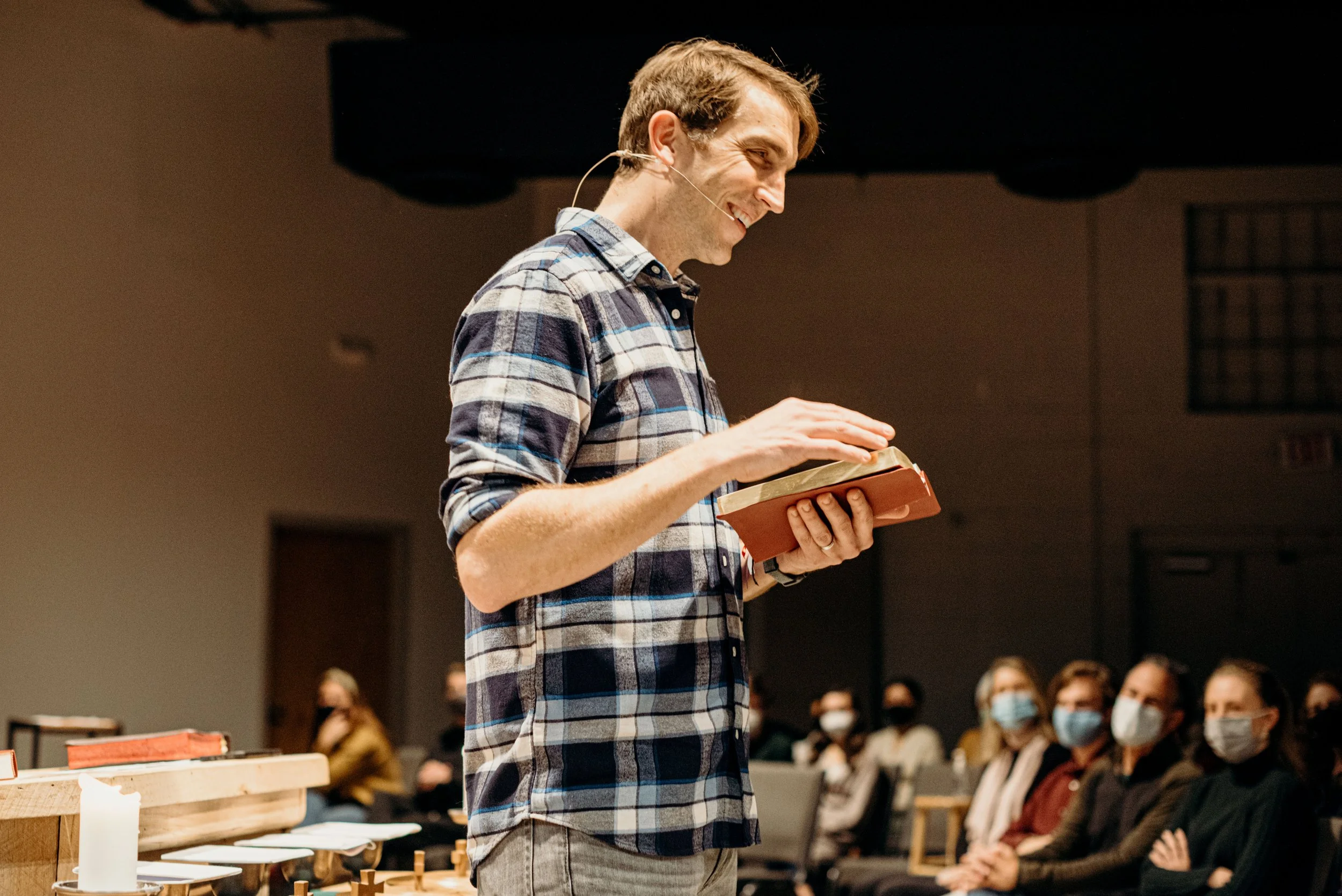 A man in a checkered shirt is smiling and reading from a book while standing in front of an audience in a lecture hall.
