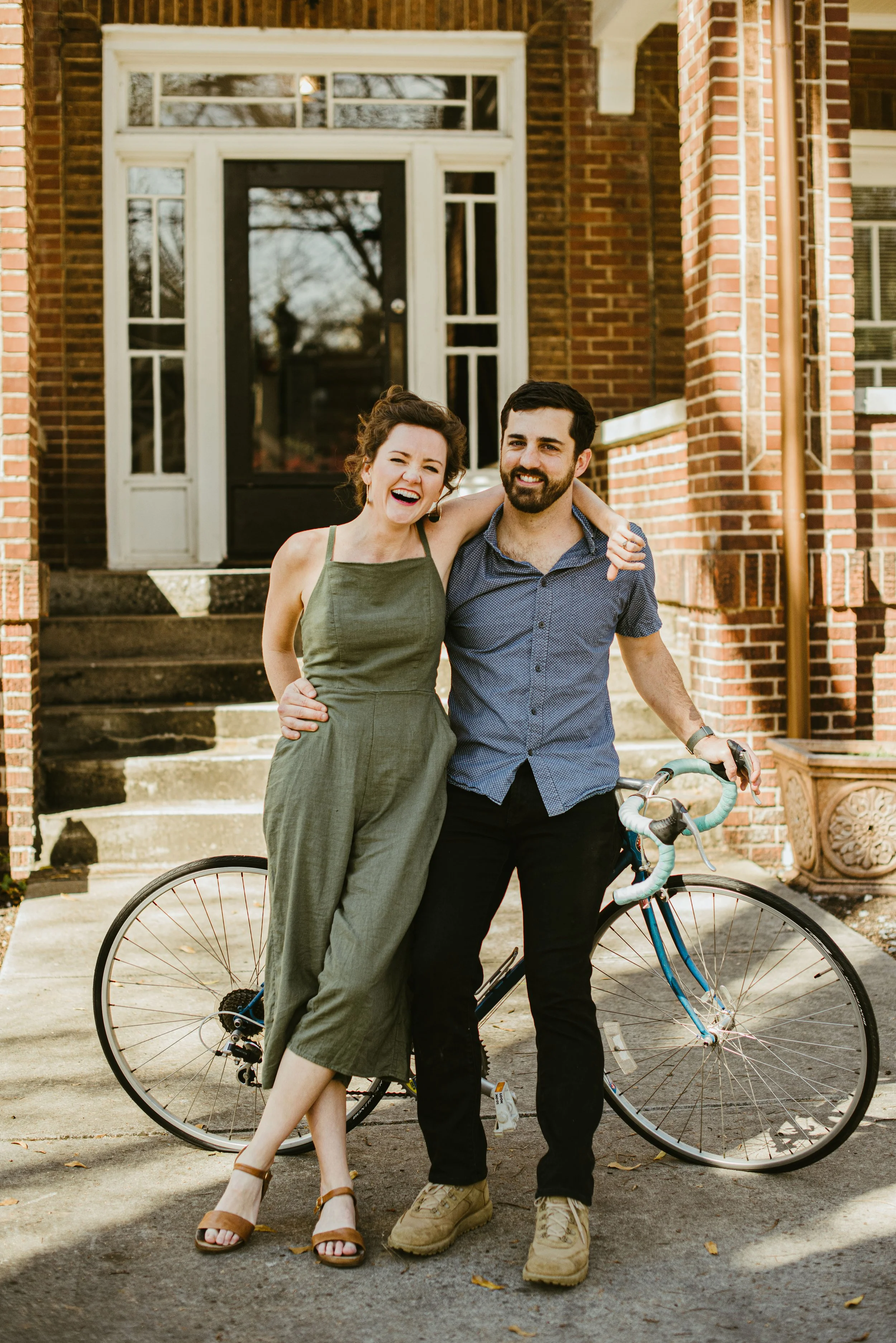 A smiling young woman and a smiling young man standing on a sidewalk, with the woman's arm around the man's shoulder, in front of a brick building. The woman is wearing a green dress and sandals, and the man is wearing a blue patterned shirt and beig