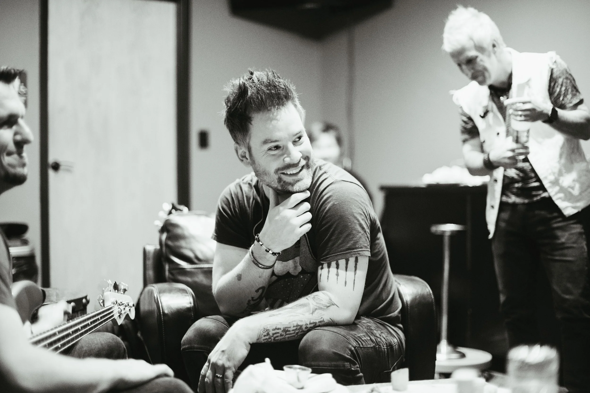 A group of people, including a man with tattoos and a woman with short blond hair, are sitting around a table enjoying a casual gathering. One person is playing guitar while others chat and smile.
