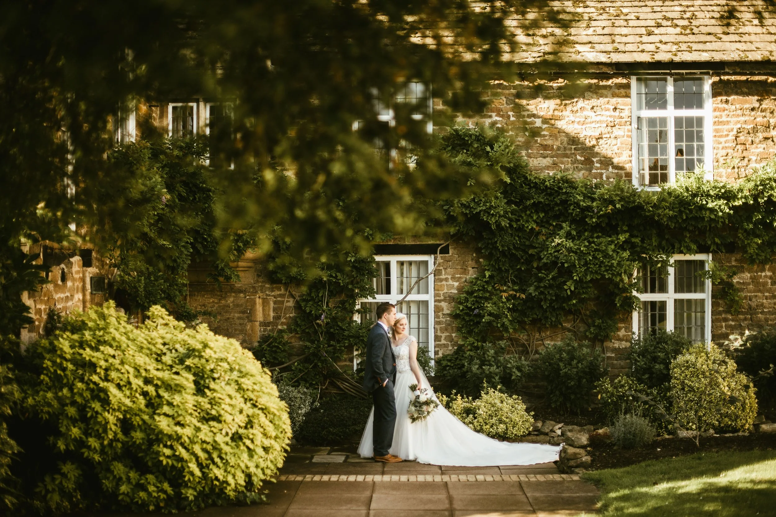 A bride and groom standing outdoors in front of a brick house covered partly with green ivy, surrounded by lush greenery and bushes, during a wedding ceremony or photo shoot.