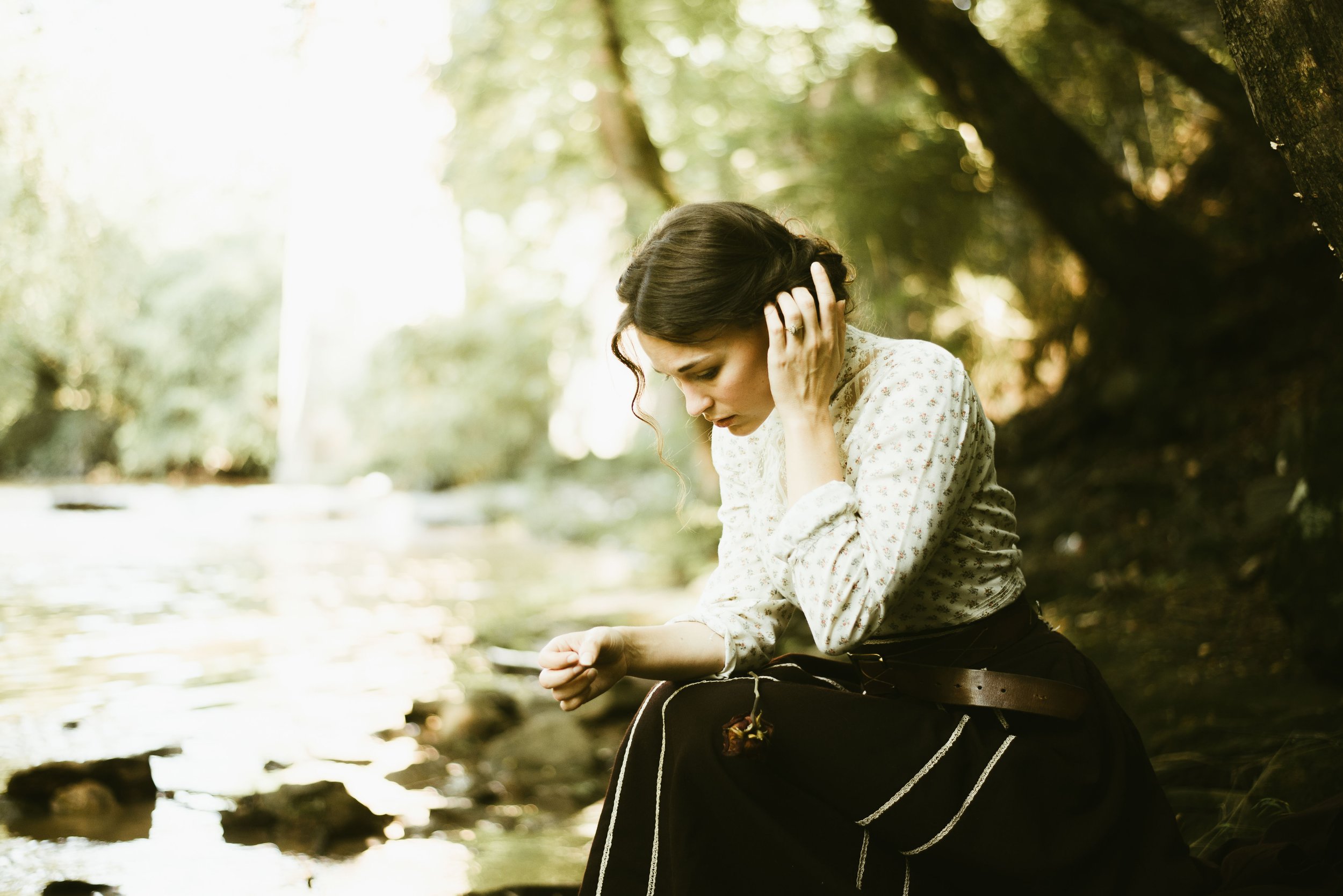 A woman sitting outdoors near a stream or river, holding her head with one hand and looking down, surrounded by trees with sunlight filtering through the leaves.