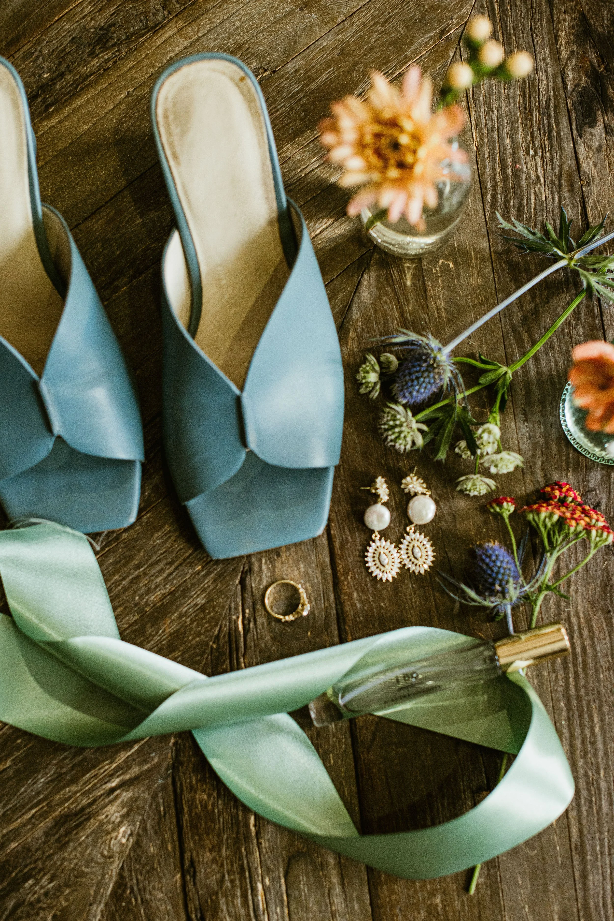 Blue high-heeled shoes with light green satin ribbons, a small bouquet of flowers, and jewelry including earrings and rings, arranged on a wooden surface.