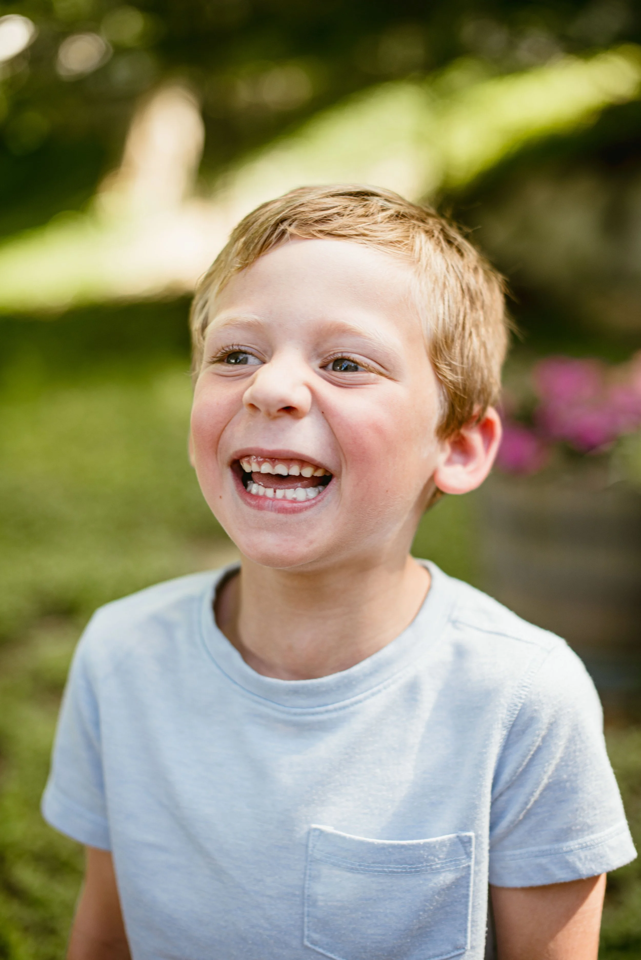 A young boy with light brown hair smiling outdoors, wearing a light blue t-shirt. The background is blurred greenery with sunlight.