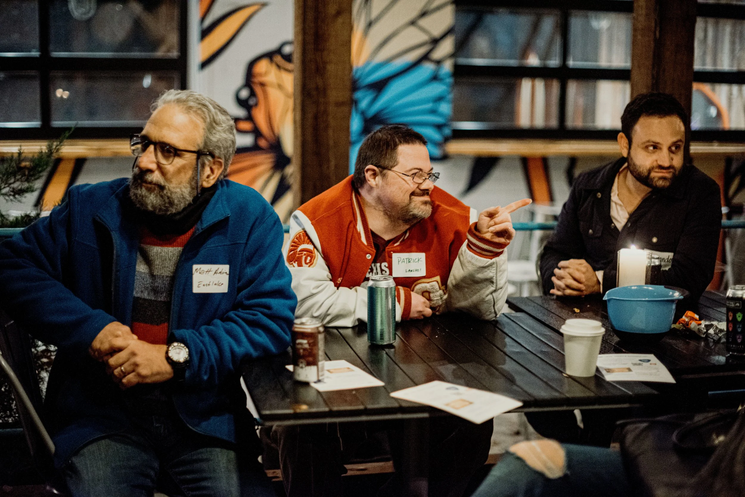 Three men sit at a table in a dimly lit indoor setting with graffiti-style art on the wall behind them. The man on the left has gray hair and a beard, wearing glasses, a blue jacket, and a name tag. The middle man, also with glasses and a beard, is w