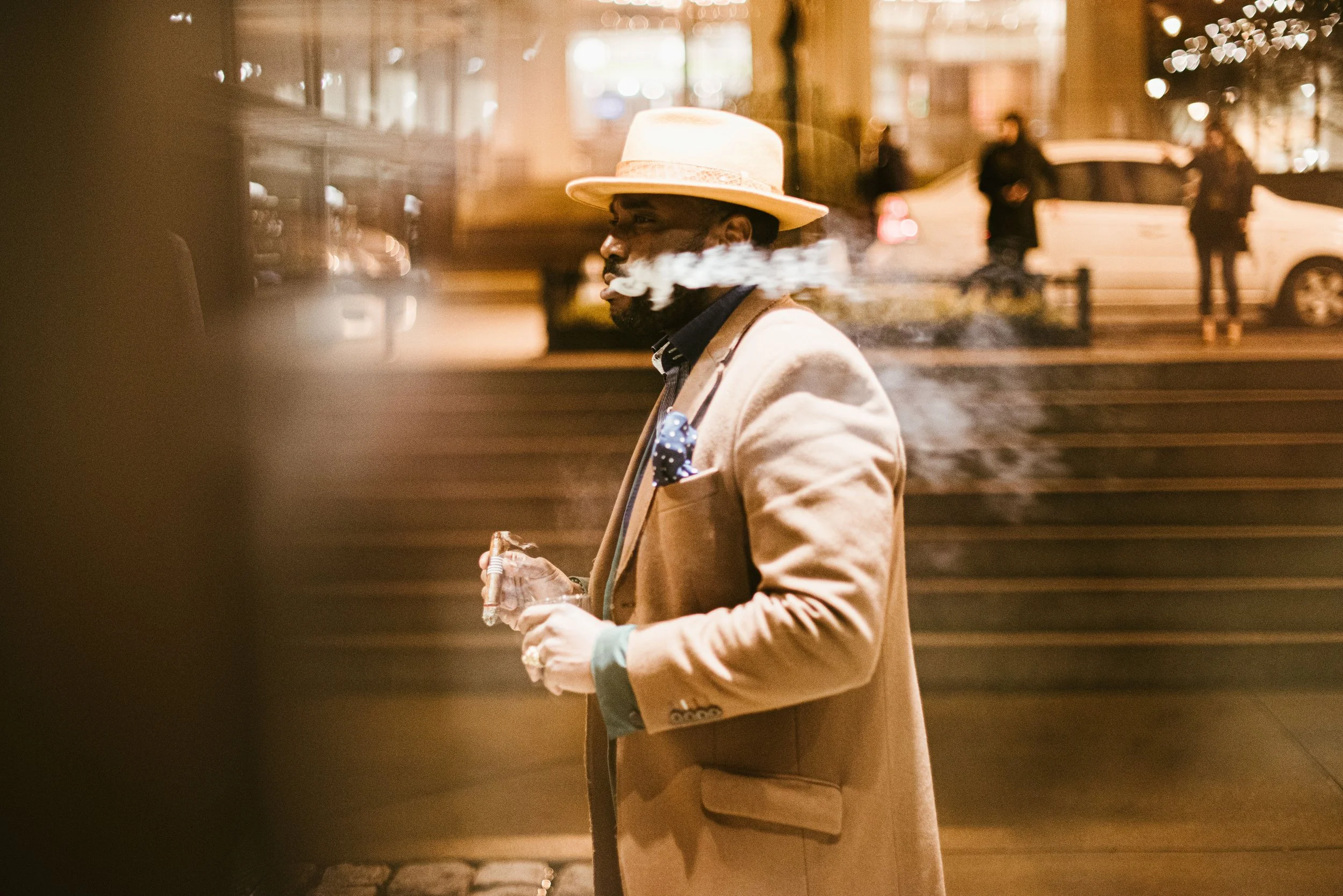 A man wearing a beige blazer, a black shirt, a polka-dotted pocket square, and a wide-brimmed hat, smoking and holding a drink in hand, standing on a city street at night with blurred pedestrians and cars in the background.