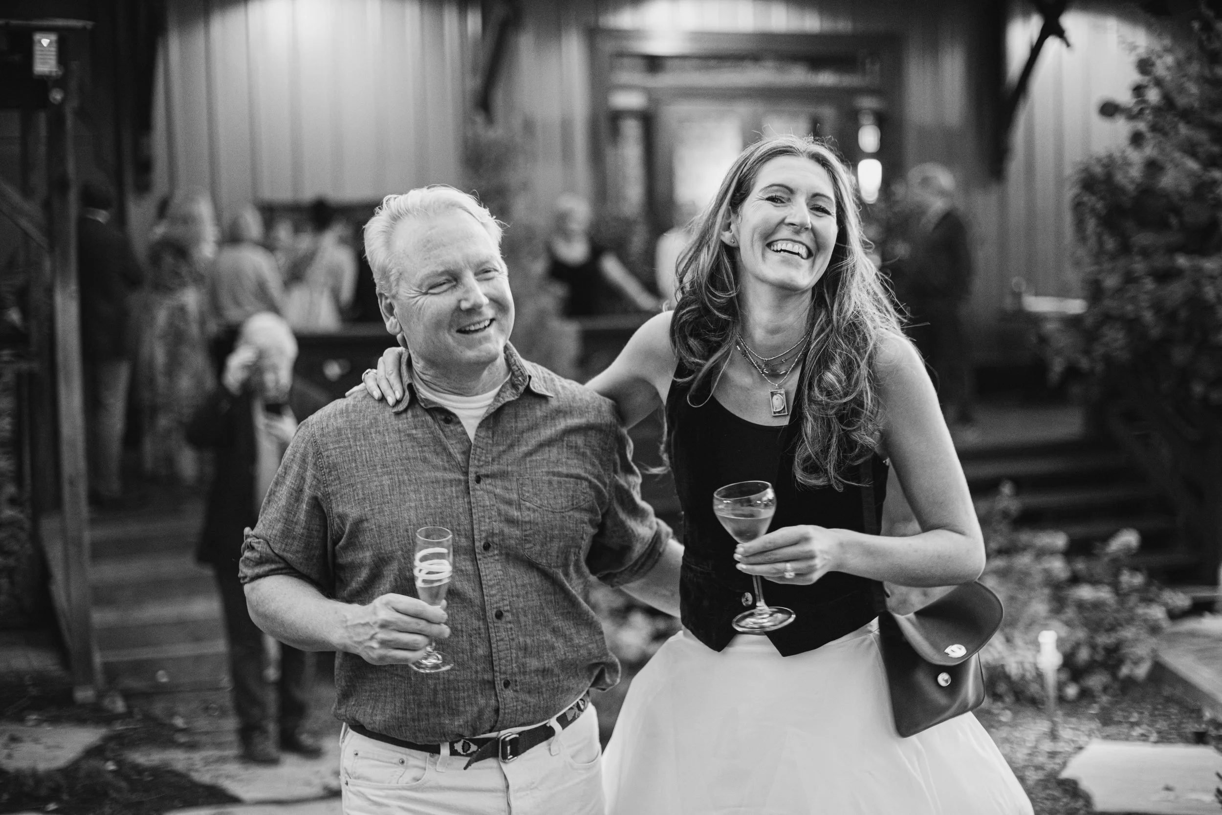 Black and white photo of a man and woman smiling and holding drinks at a social gathering. The man has light-colored hair, and the woman has long, wavy hair and is wearing layered necklaces and a black top. The background shows other people and a rus
