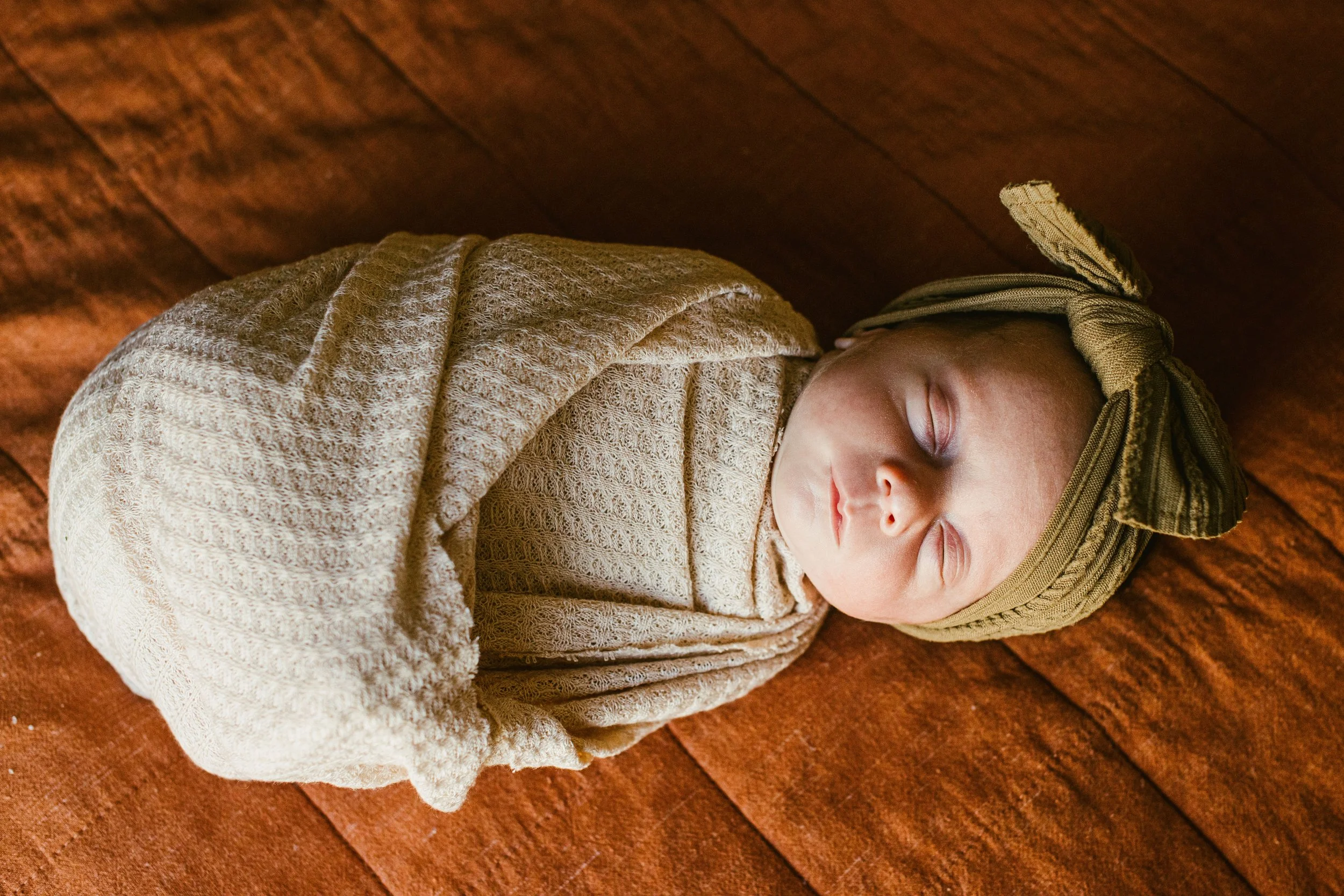 A young baby with a tan hat and beige textured blanket, sleeping on a reddish-brown wooden floor.