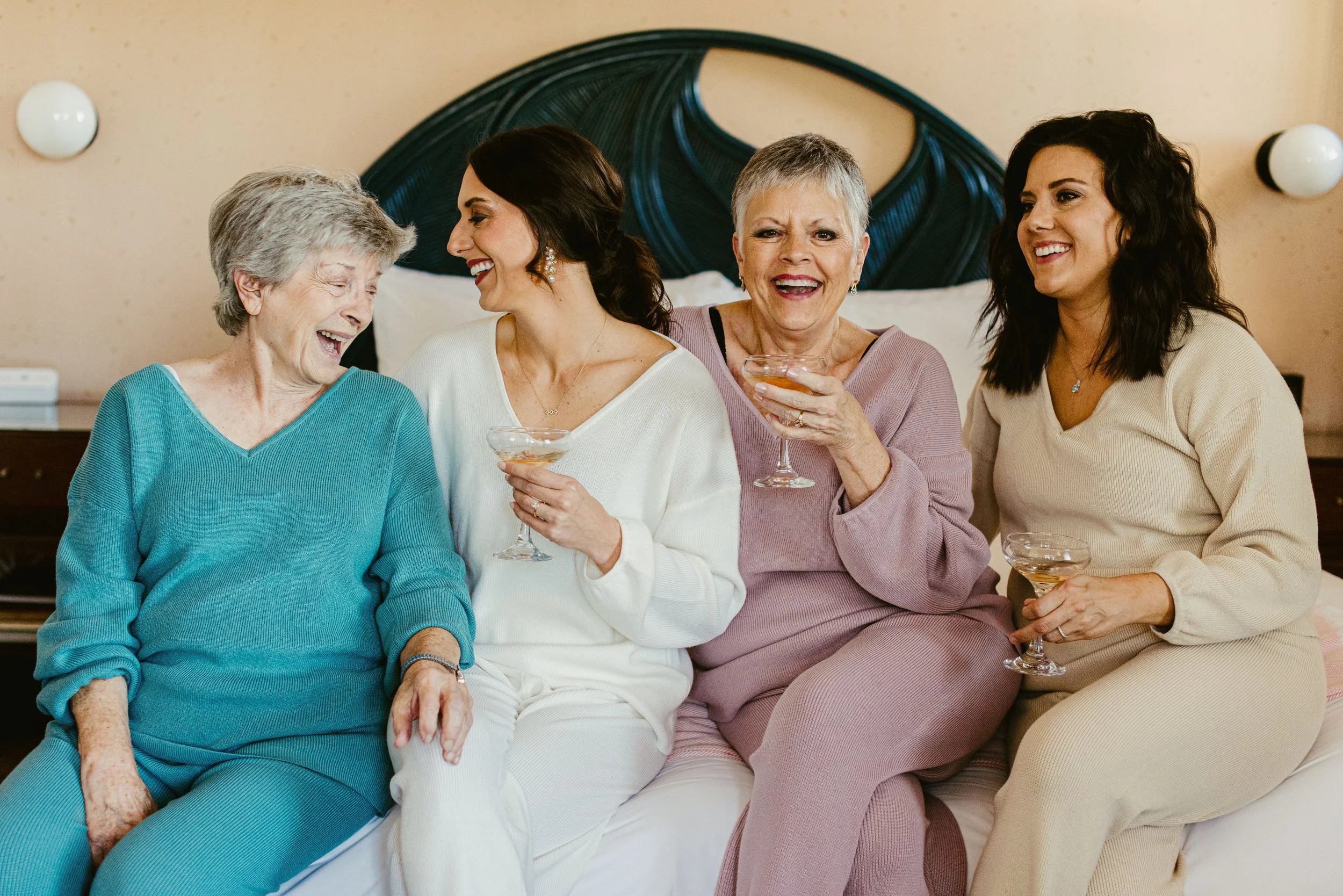 Five women sitting on a bed, laughing and holding drinks, in a warmly decorated room.