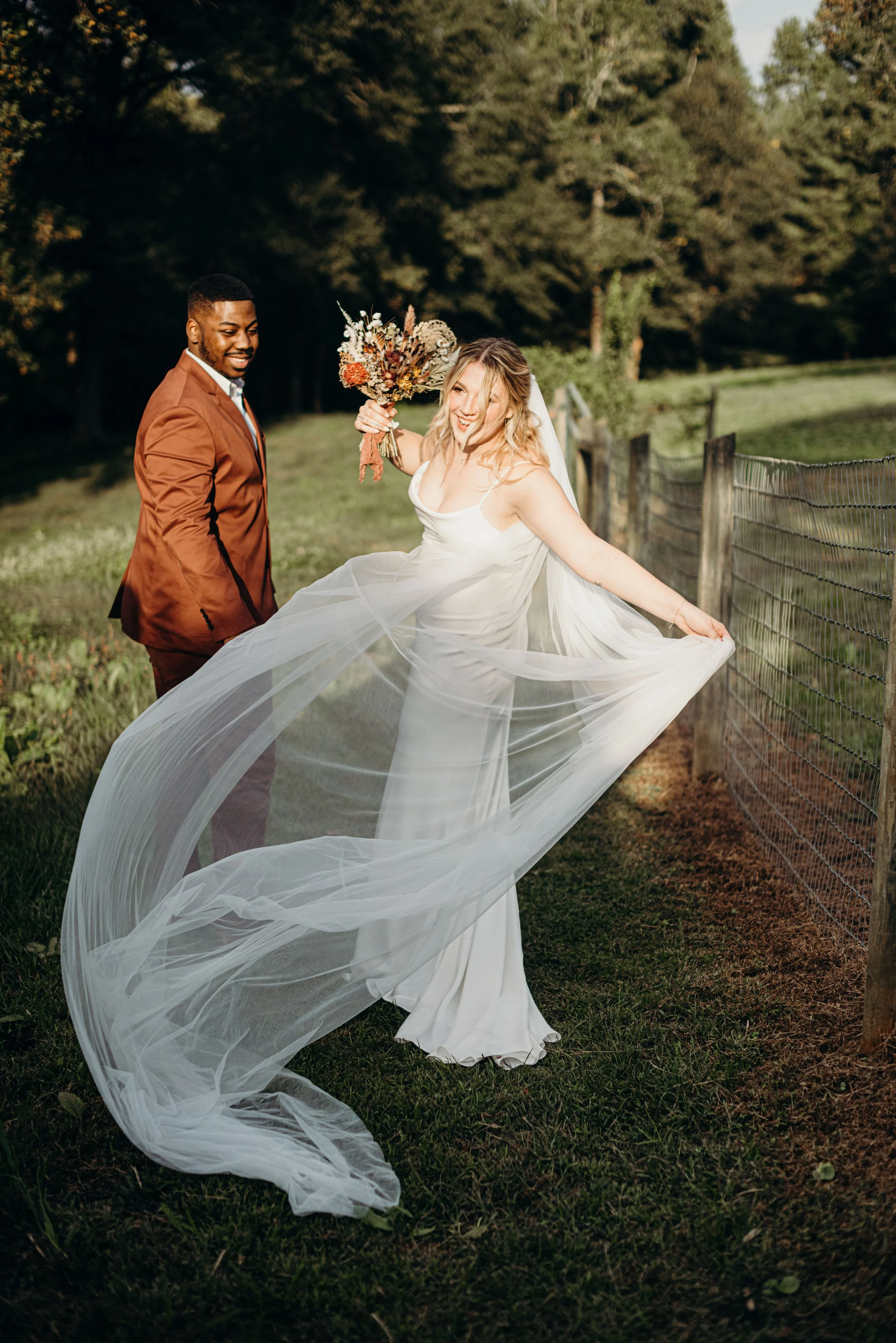 A bride and groom standing outdoors on a grassy area near a wooden fence, with the bride playfully holding out her flowing wedding dress and the groom smiling at her, surrounded by trees.