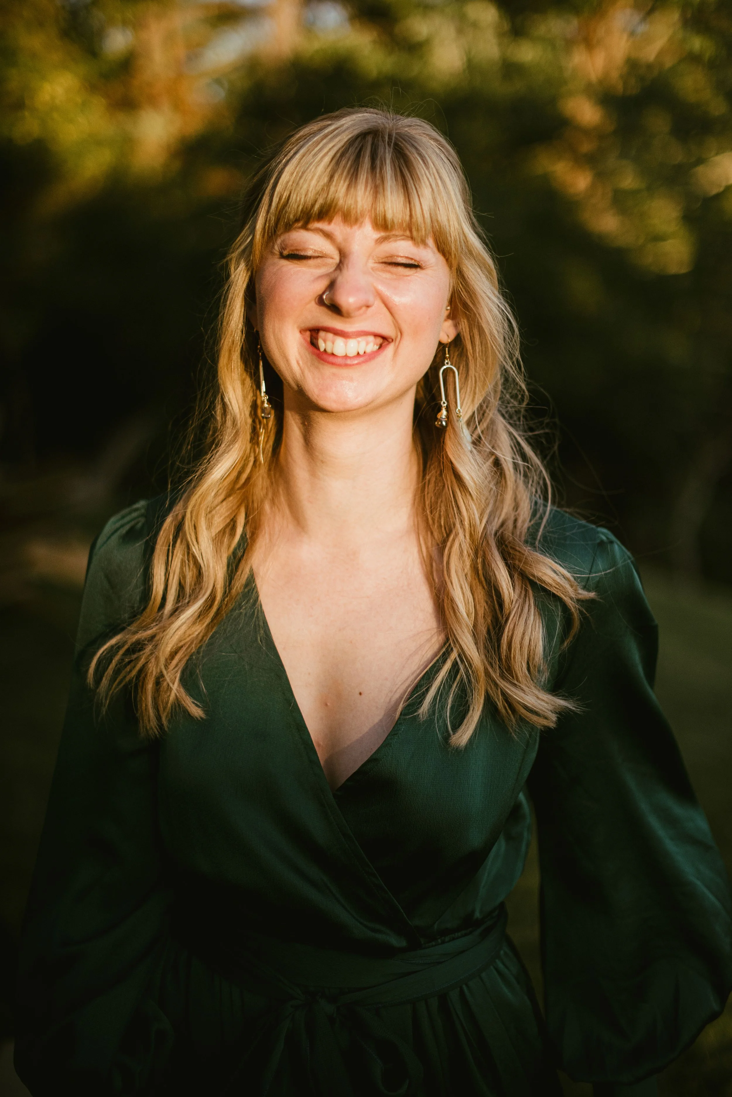A woman with blonde hair and bangs smiling with her eyes closed outdoors during daytime, wearing earrings and a dark green top.