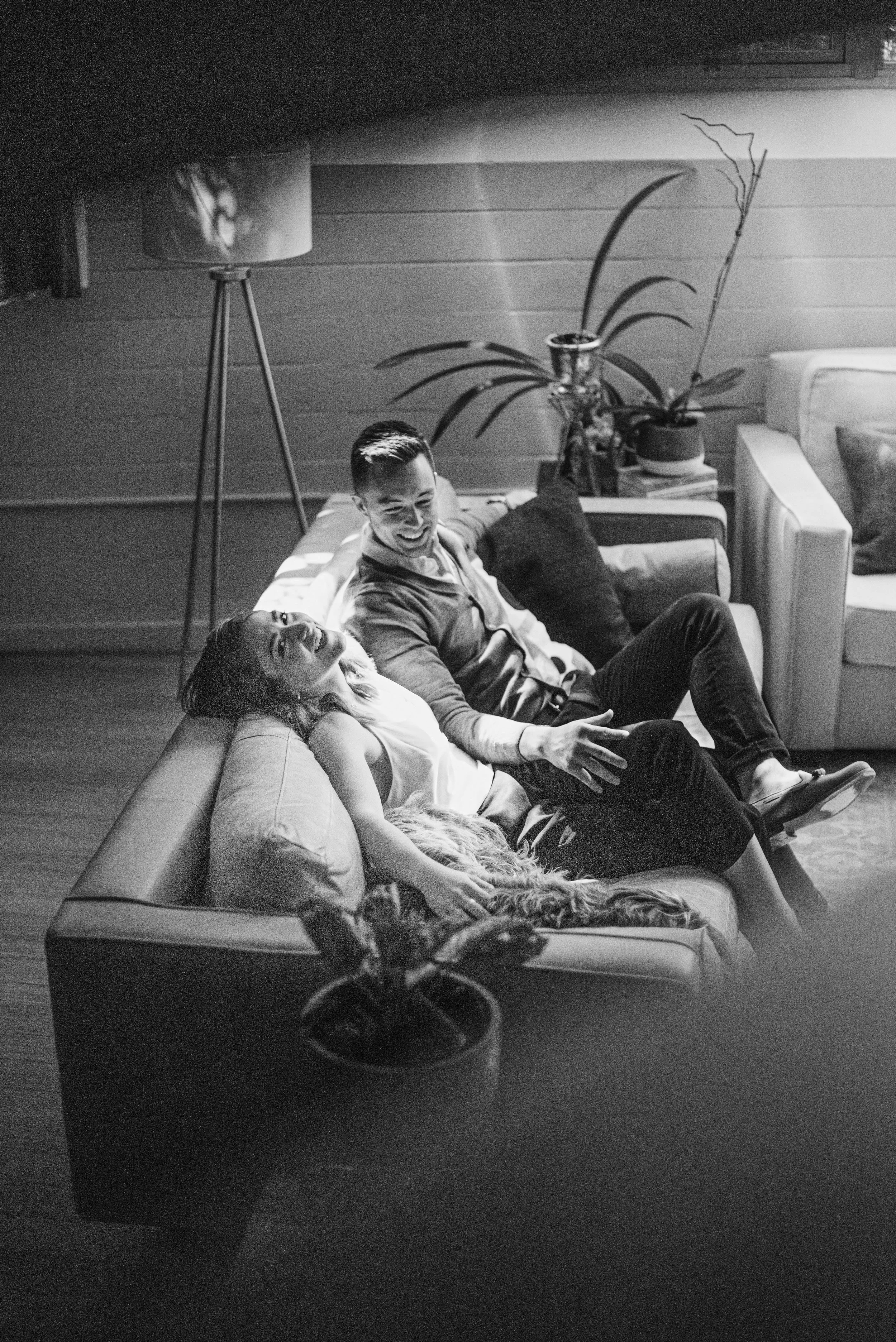 A black and white photo of three people relaxing on a sofa in a living room. Two women are lying down, and a man is sitting up, all smiling and engaging with each other. There is a tall floor lamp and a large plant on a side table behind them.