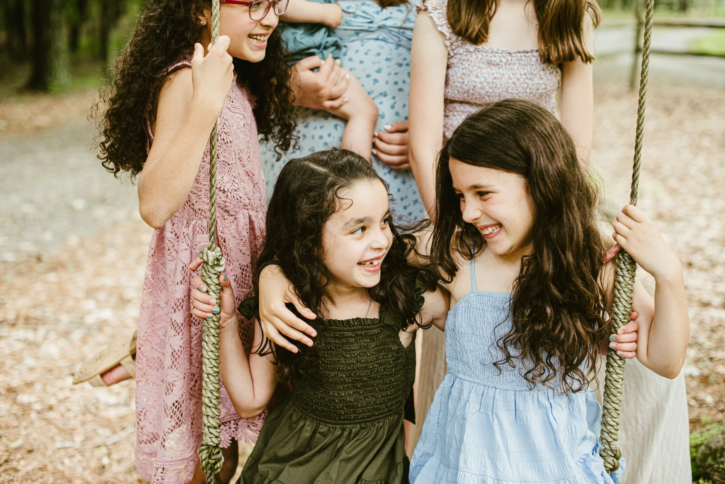 Four children, three girls and one boy, playing and laughing on a swing set outdoors. The girls are smiling and talking, holding the swing ropes, and the boy is sitting on the swing. They are in a wooded area with a blurred background.