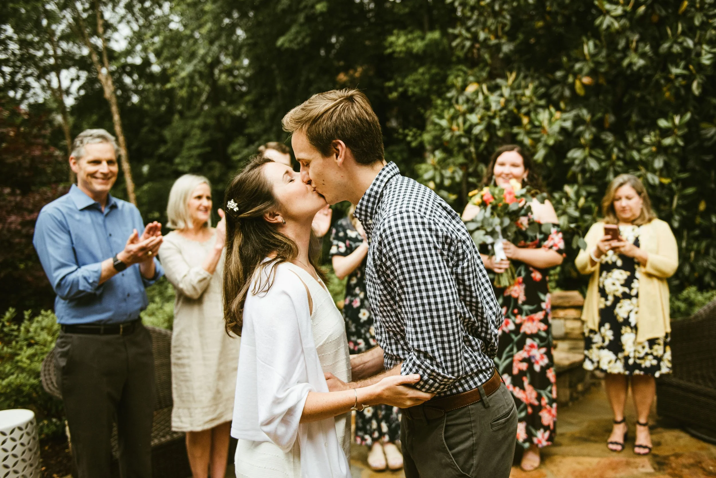 A group of people gathered outdoors in a garden, with a couple kissing in the foreground. The woman is wearing a white dress, and the man is wearing a checkered shirt. Several people are clapping and smiling, and a woman is holding a bouquet of flowe