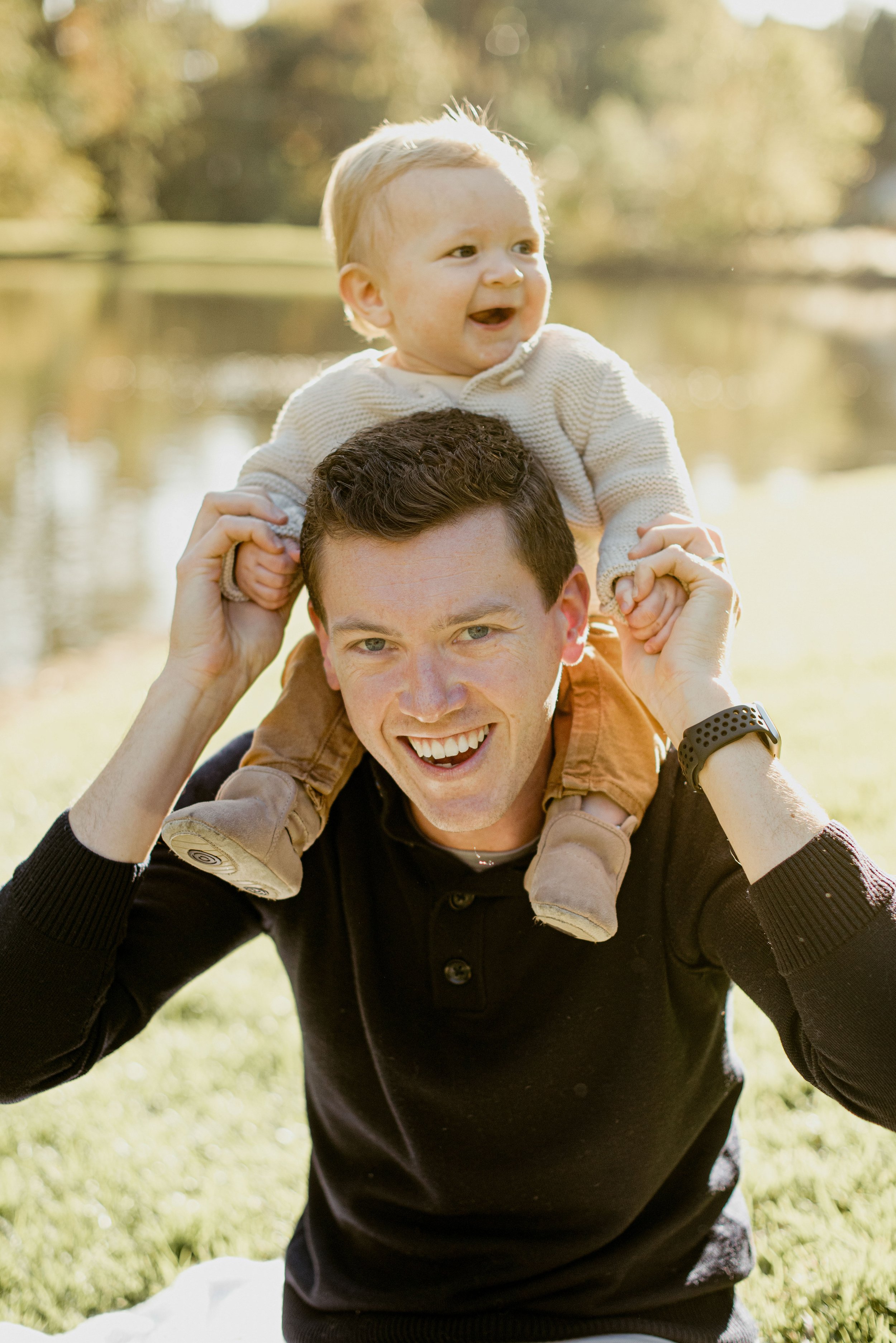 A man carrying a young child on his shoulders outdoors during daytime, with a blurred background of grass, trees, and a body of water, indicative of a park.