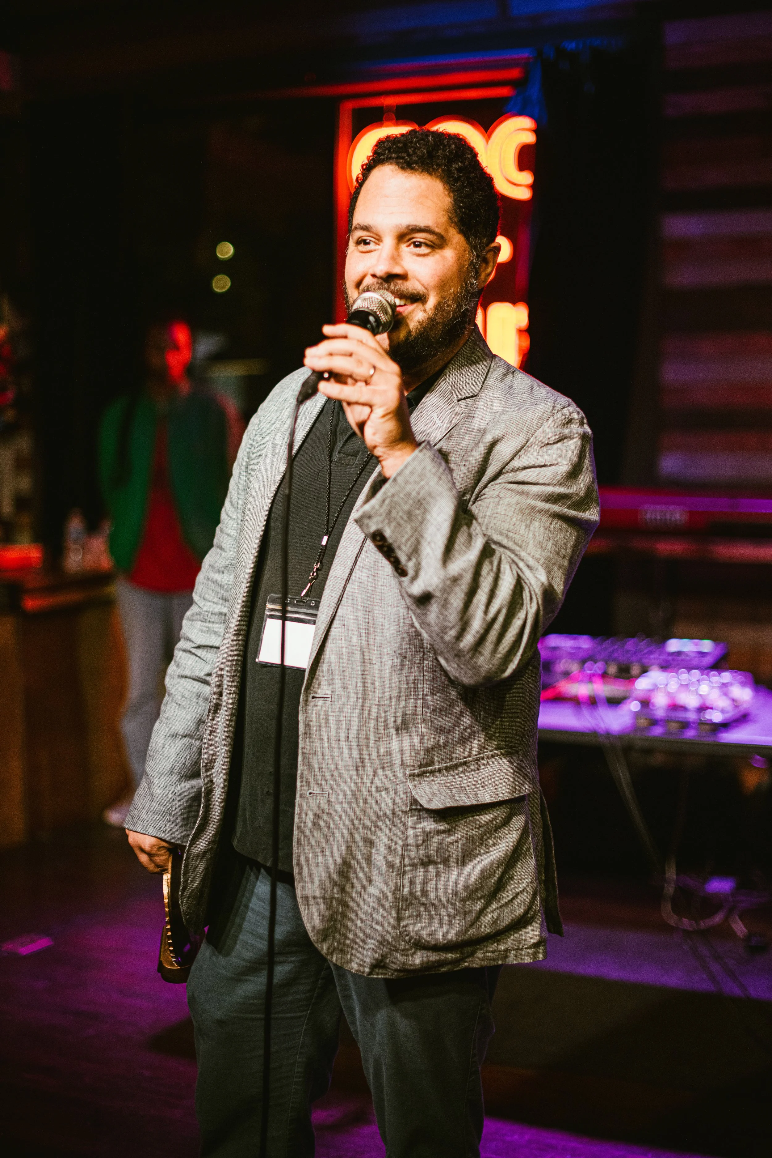 A man in a gray blazer and black shirt holding a microphone, standing in a dimly lit setting with a neon sign in the background, possibly performing or speaking at an event.
