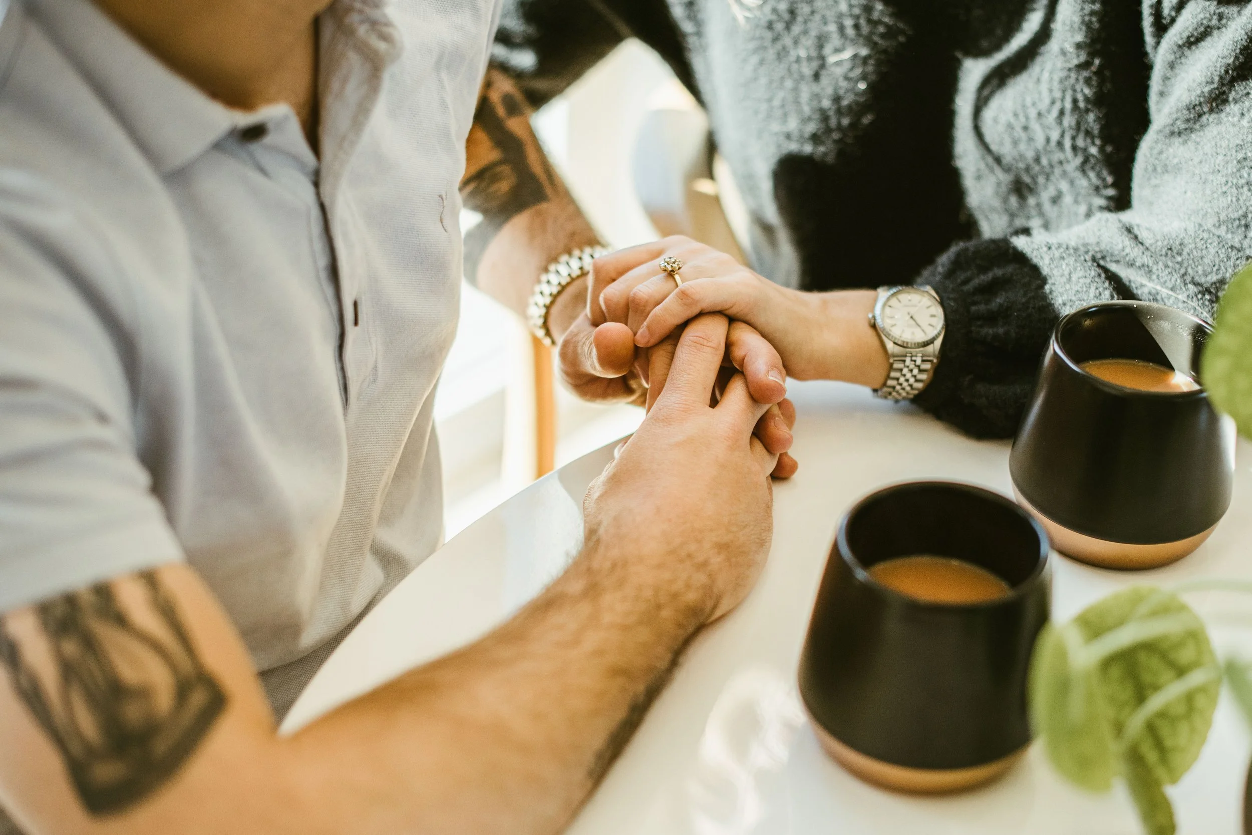 Two people holding hands on a white table with black cups of coffee, some plants and cups in the background.