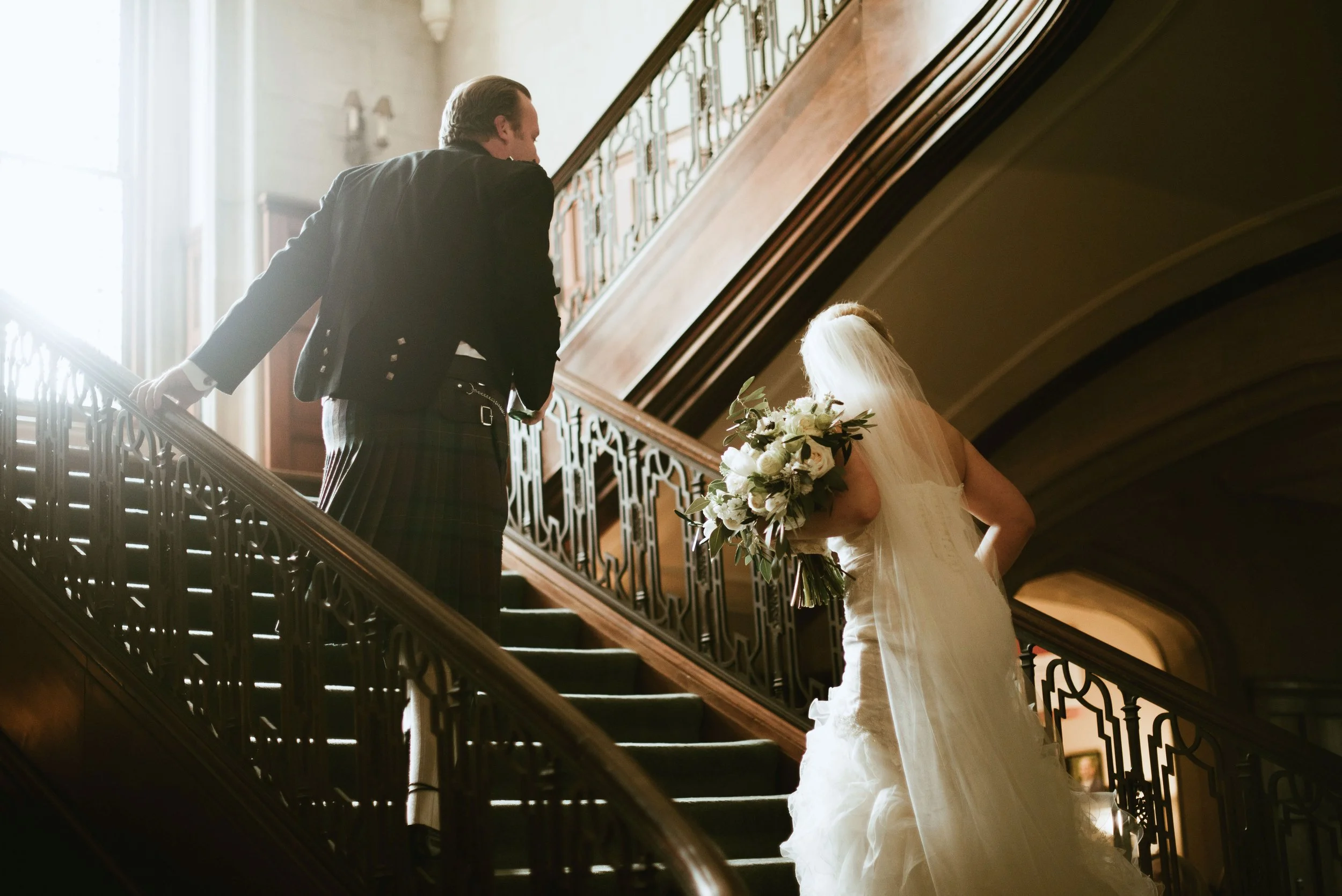 A bride holding a bouquet of white flowers, standing on a staircase, looking up at a man in formal attire, possibly at a wedding ceremony in a grand building.