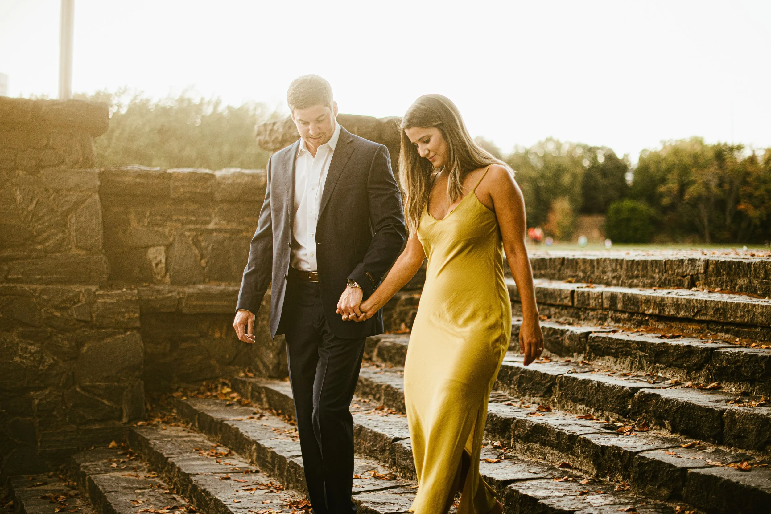 A man and a woman walking hand-in-hand outdoors during sunset, with a stone wall and autumn trees in the background.