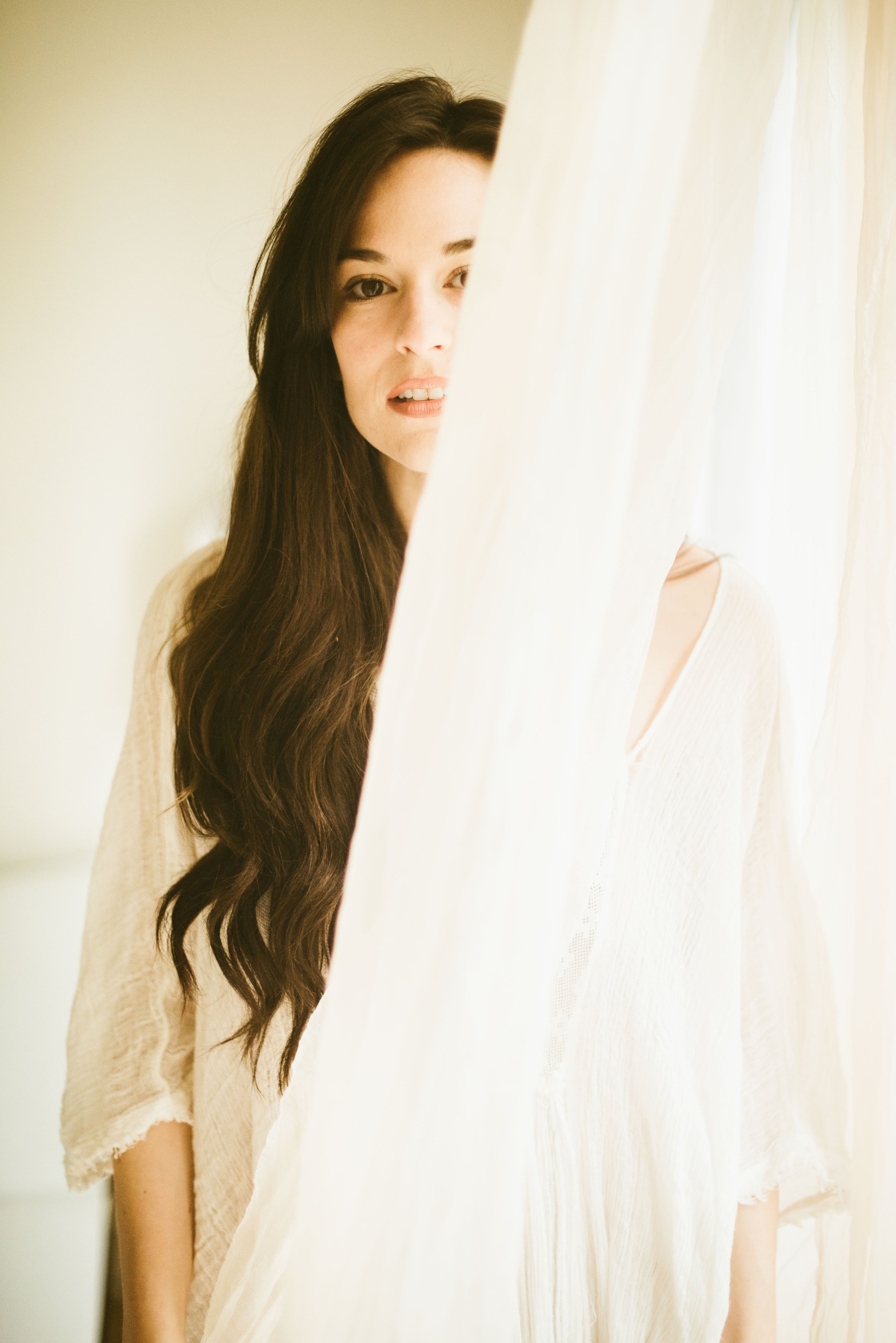 A woman with long dark hair standing behind a white curtain, peeking out from behind it.