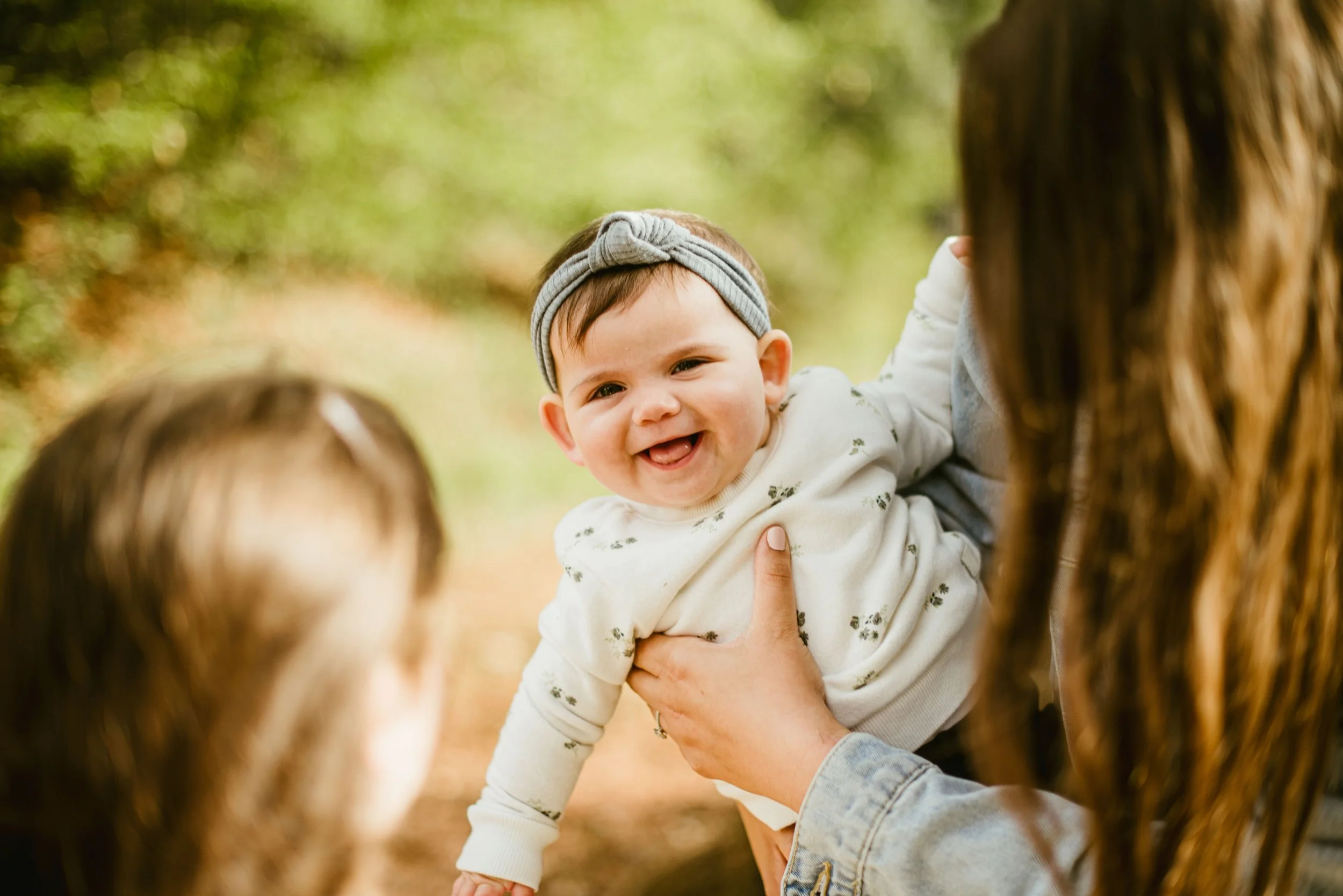 A smiling baby with a headband being held up by a woman outdoors among trees.