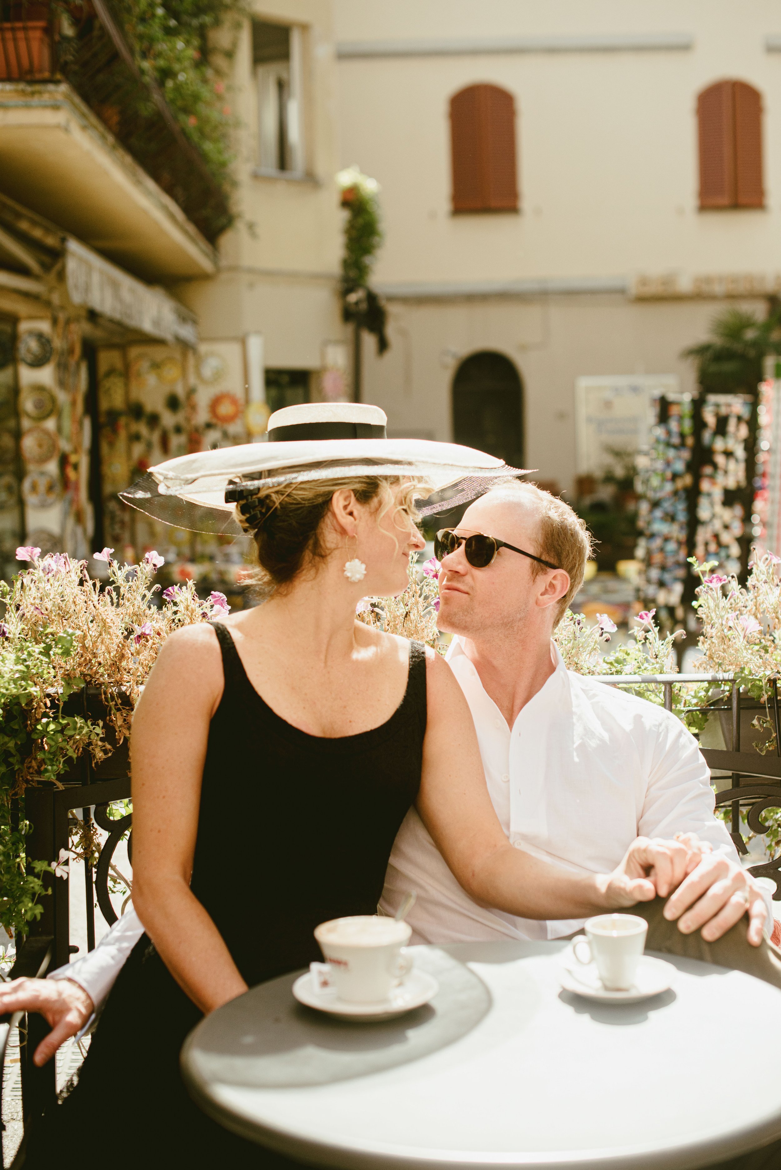 A man and a woman sit closely together at an outdoor café table, with the woman wearing a wide-brimmed hat and the man wearing sunglasses. They are looking at each other, surrounded by flowers and a building with closed shutters in the background.