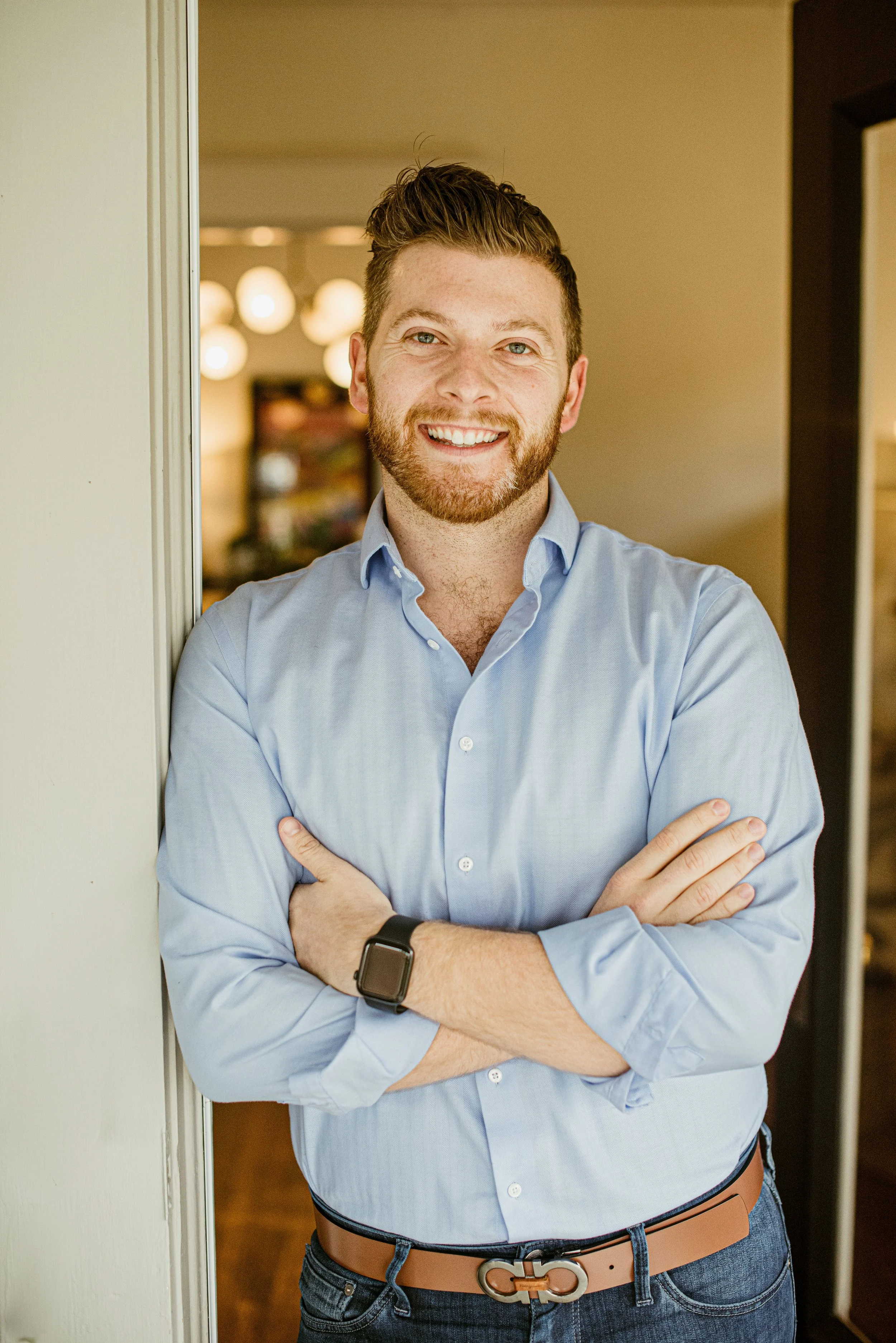 A man with a beard and short, styled hair is standing with arms crossed, smiling at the camera, wearing a light blue dress shirt, a watch, and a belt with a decorative buckle, indoors with warm lighting.