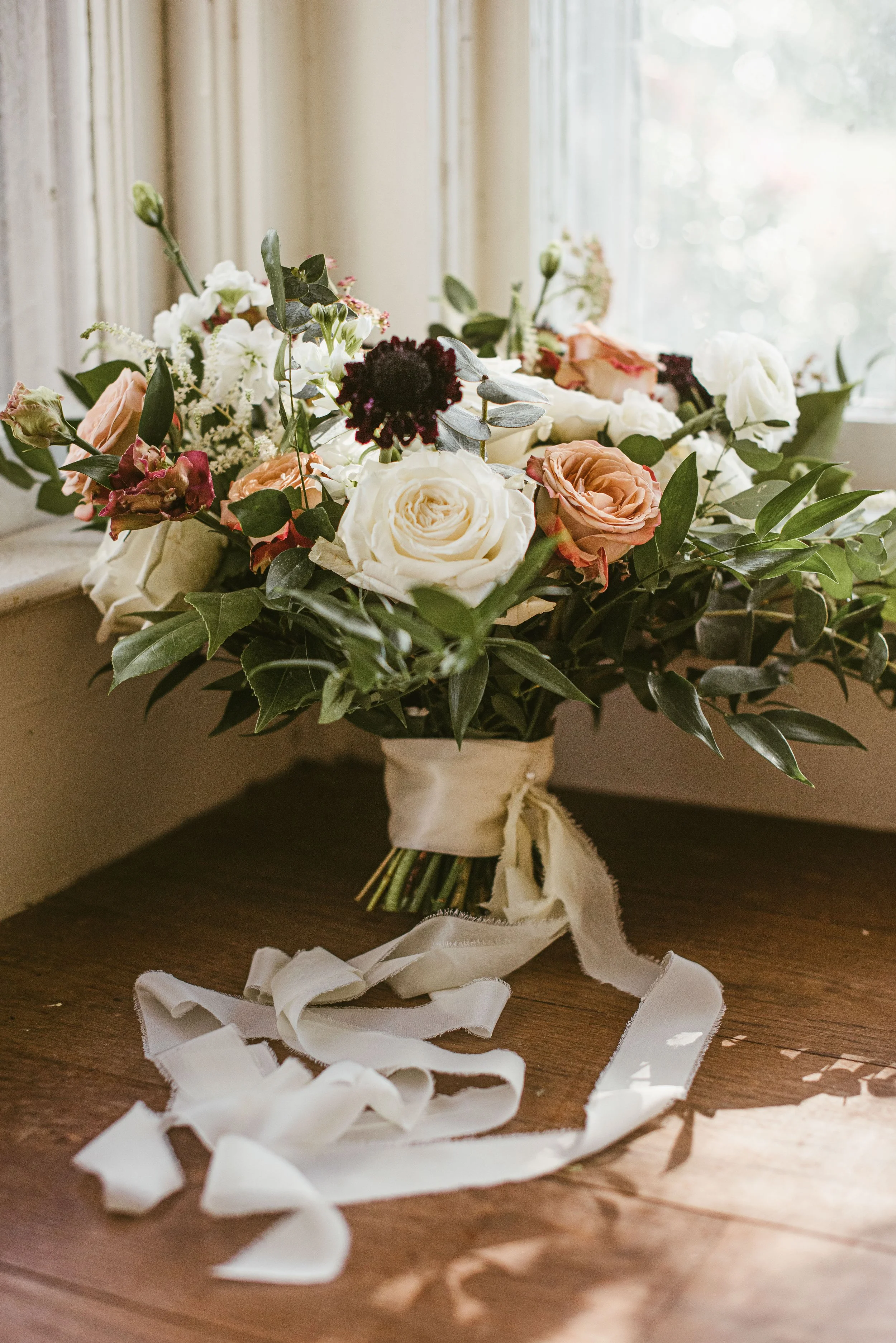 A bouquet of mixed flowers, including white roses, peaches, dark red, and white flowers, with green leaves, tied with a cream-colored ribbon, placed on a wooden surface near a window with sheer curtains.